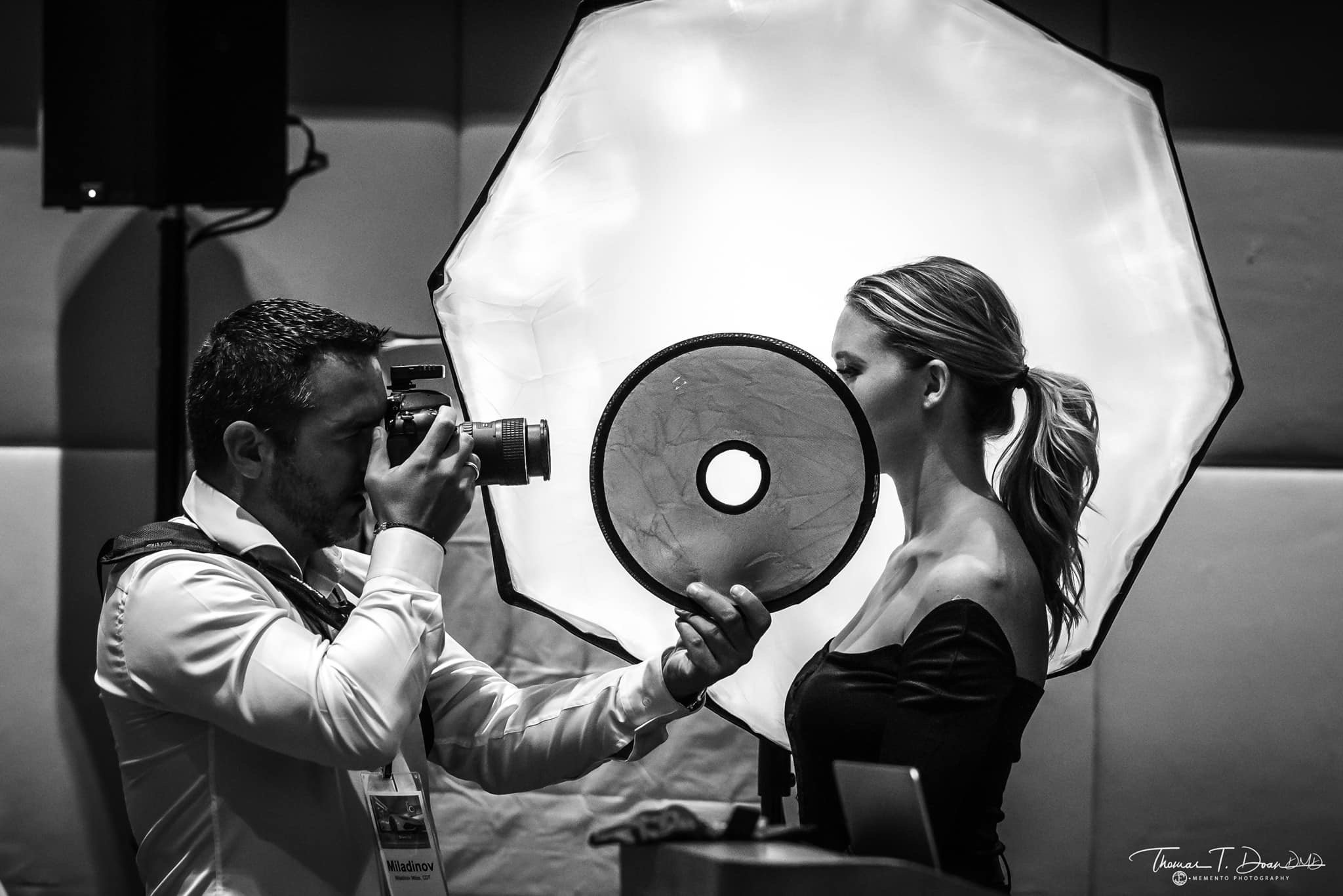 Black and white photo of a photographer taking a portrait of a woman with a studio flash umbrella. The woman is holding a reflector, and both are looking at each other during the photoshoot.