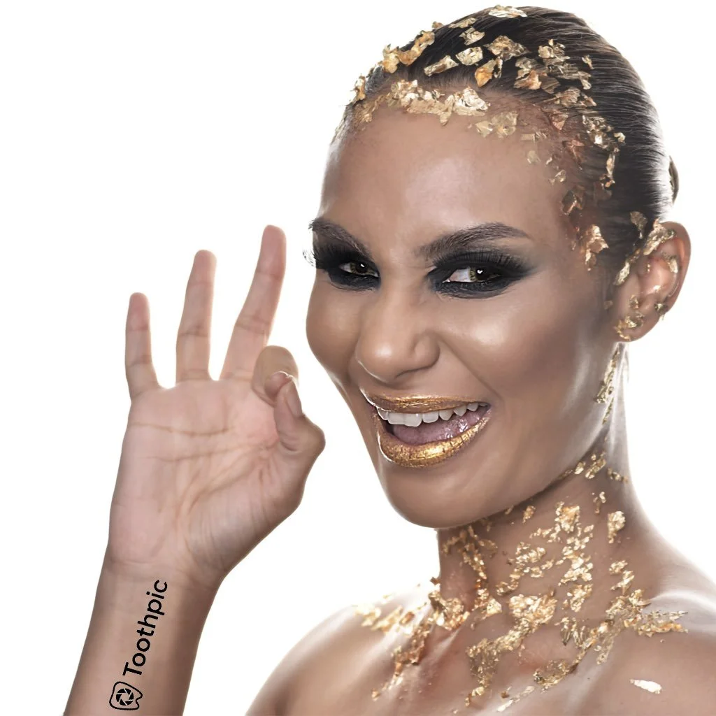 A woman with gold leaf on her hair, face, neck, and body, smiling and making an OK gesture with her hand. She has dark eye makeup and gold lipstick.
