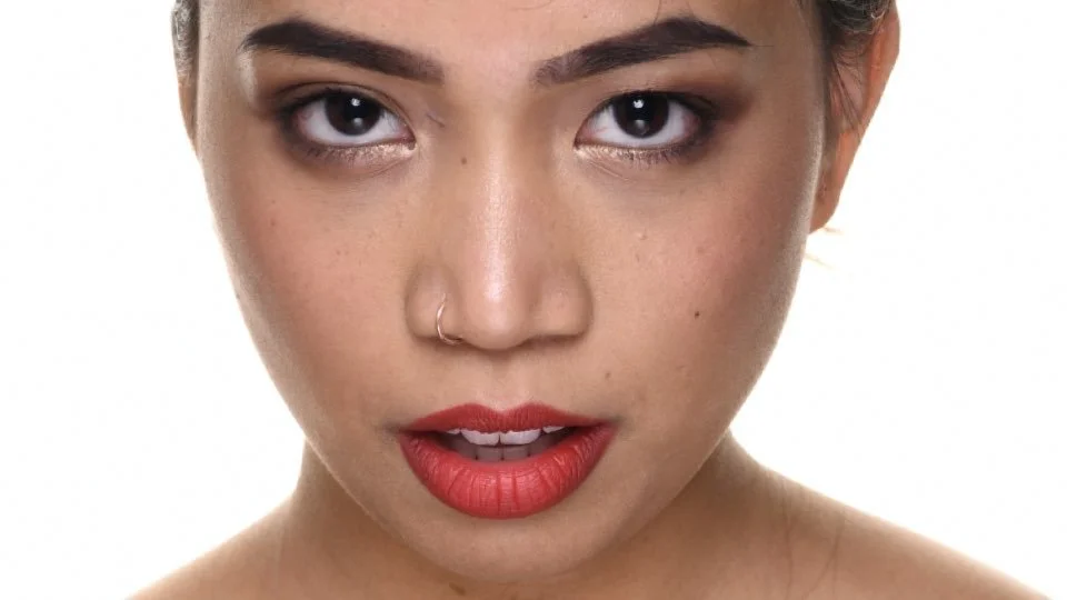 A woman with dark eyes, a nose piercing, and red lipstick looking directly at the camera with a serious expression.