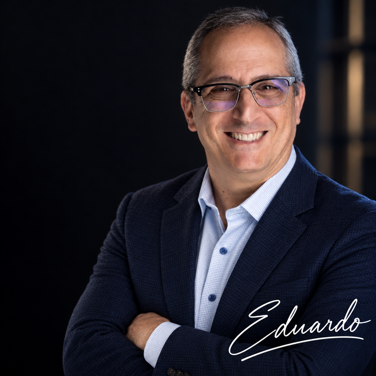 Headshot photo of a smiling man with gray hair and glasses, wearing a navy blazer and light blue shirt, crossed arms, dark background, with the name 'Eduardo' written in cursive.
