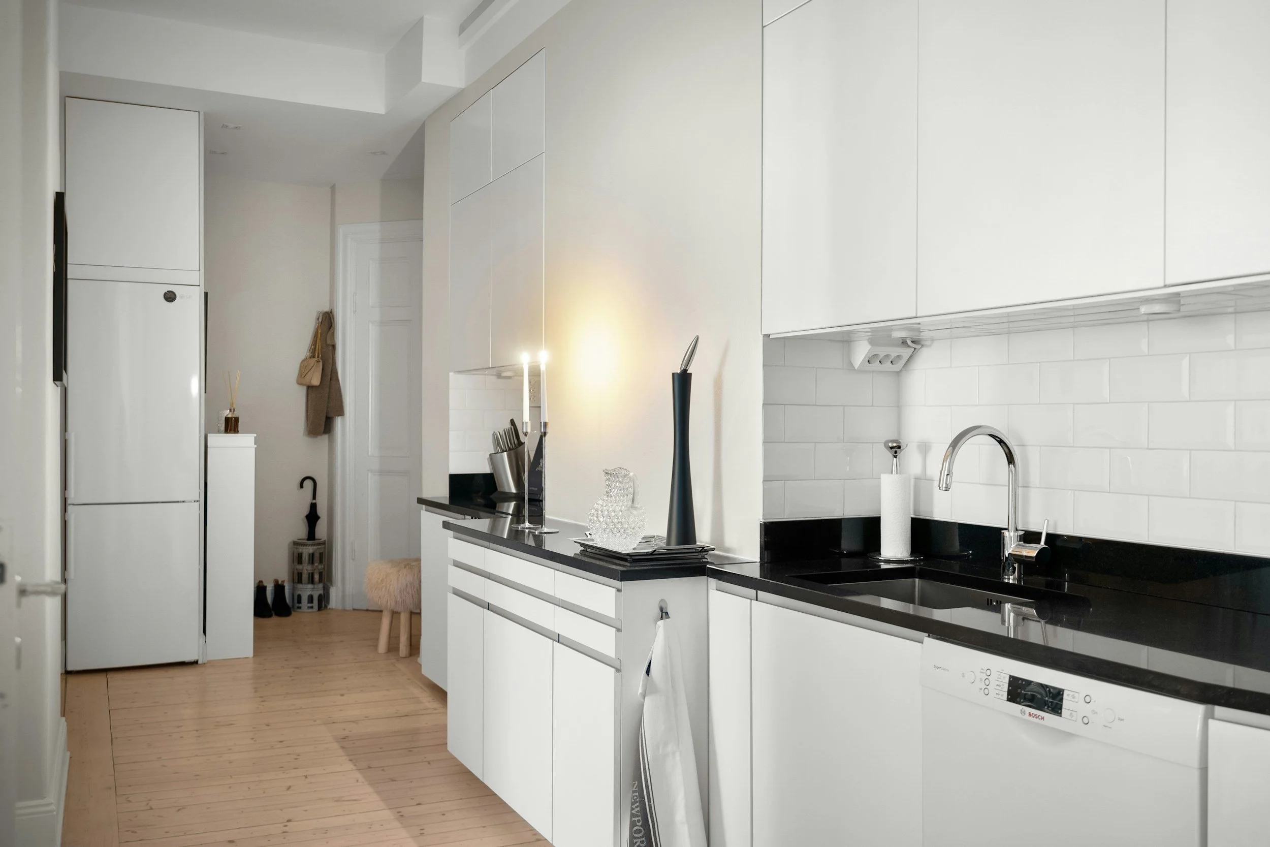 Modern, minimalist kitchen with white cabinets, black countertops, stainless steel sink, white tile backsplash, wooden floor, and kitchen appliances.