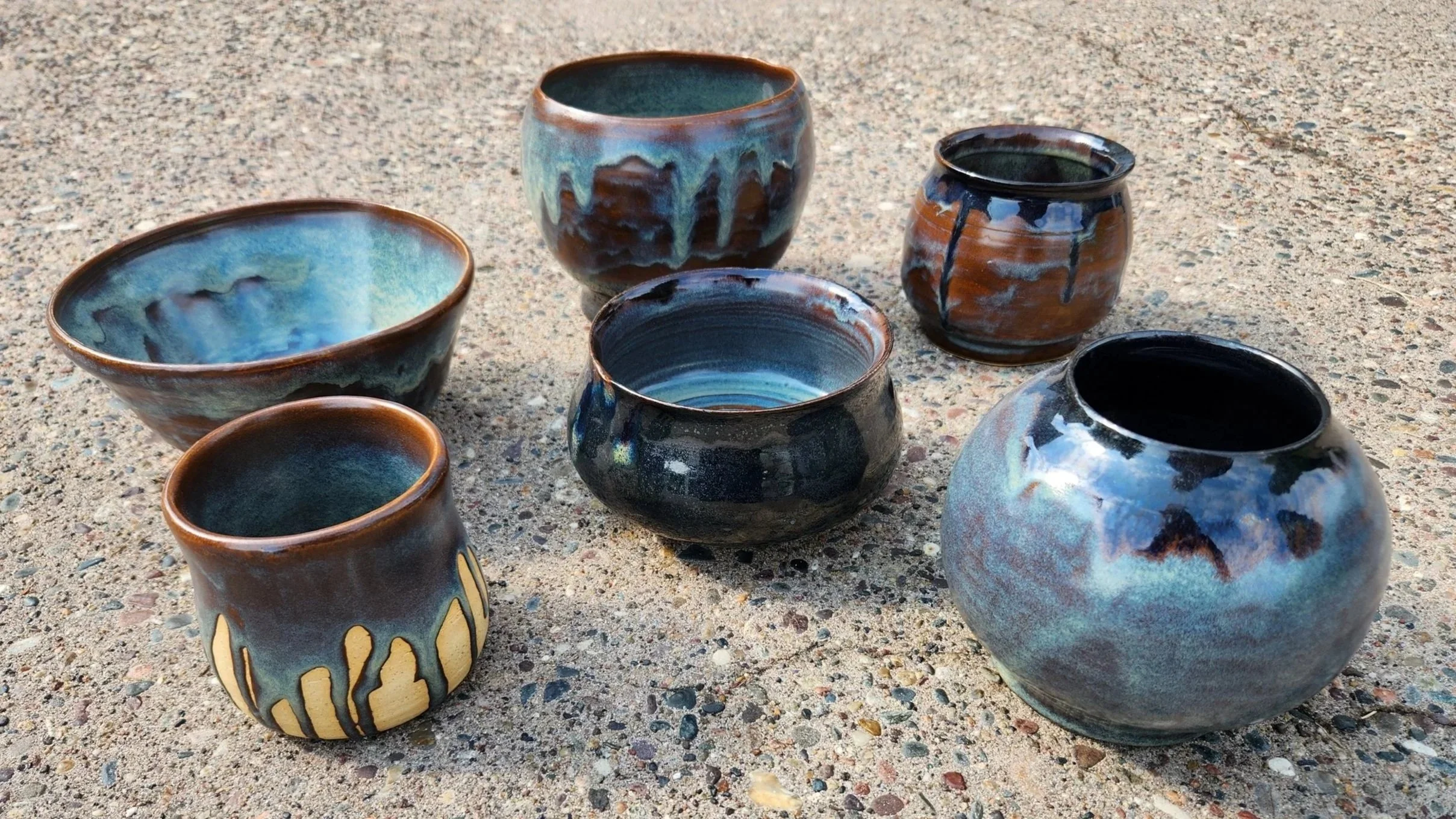 A collection of seven glazed ceramic bowls and cups on a textured concrete surface, each with a unique blue, black, and brown color pattern.