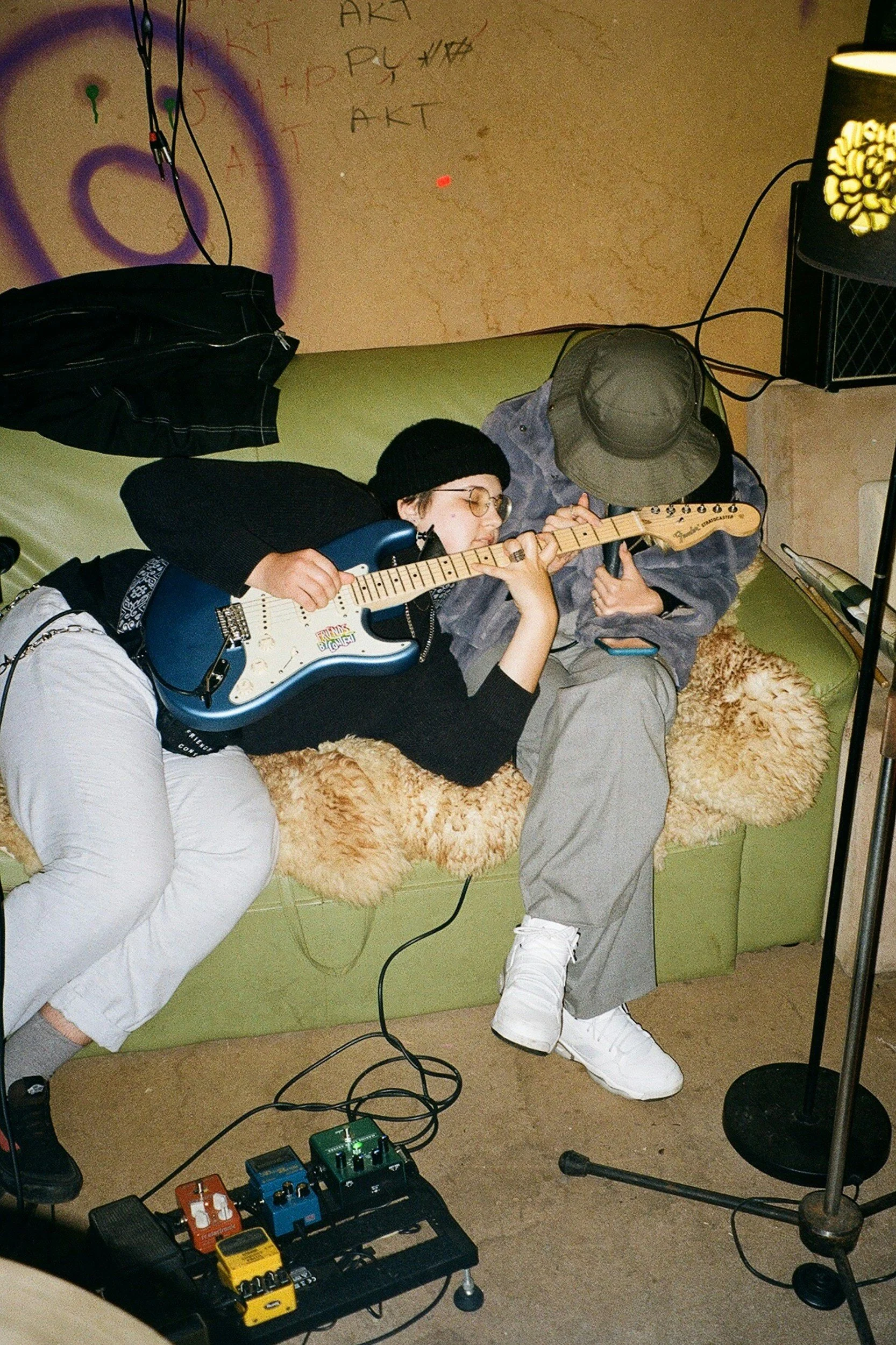 Two people are lying on a green couch, playing guitars, with a guitar pedalboard on the floor in front of them. One person is wearing a black beanie and glasses, and the other is wearing a gray cap and hoodie. A black jacket is hanging on the back of the couch. There is a wall behind them with graffiti and writings, including the word 'AKT'. A lamp with a floral design is positioned to the right, with wires and a speaker nearby.