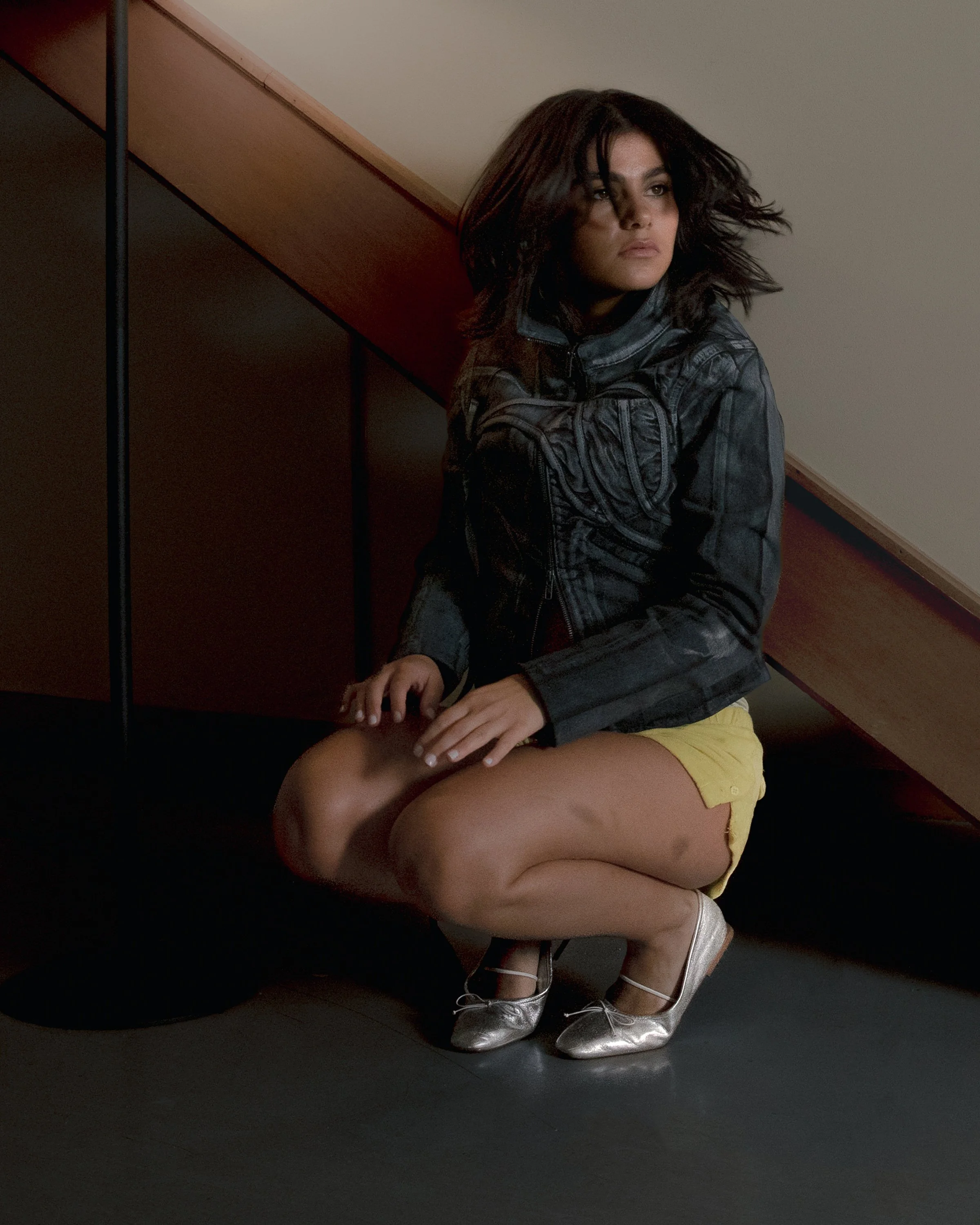 Ethel, an olive-skinned artist with dark wavy hair, crouches by a staircase in a denim jacket under amber light