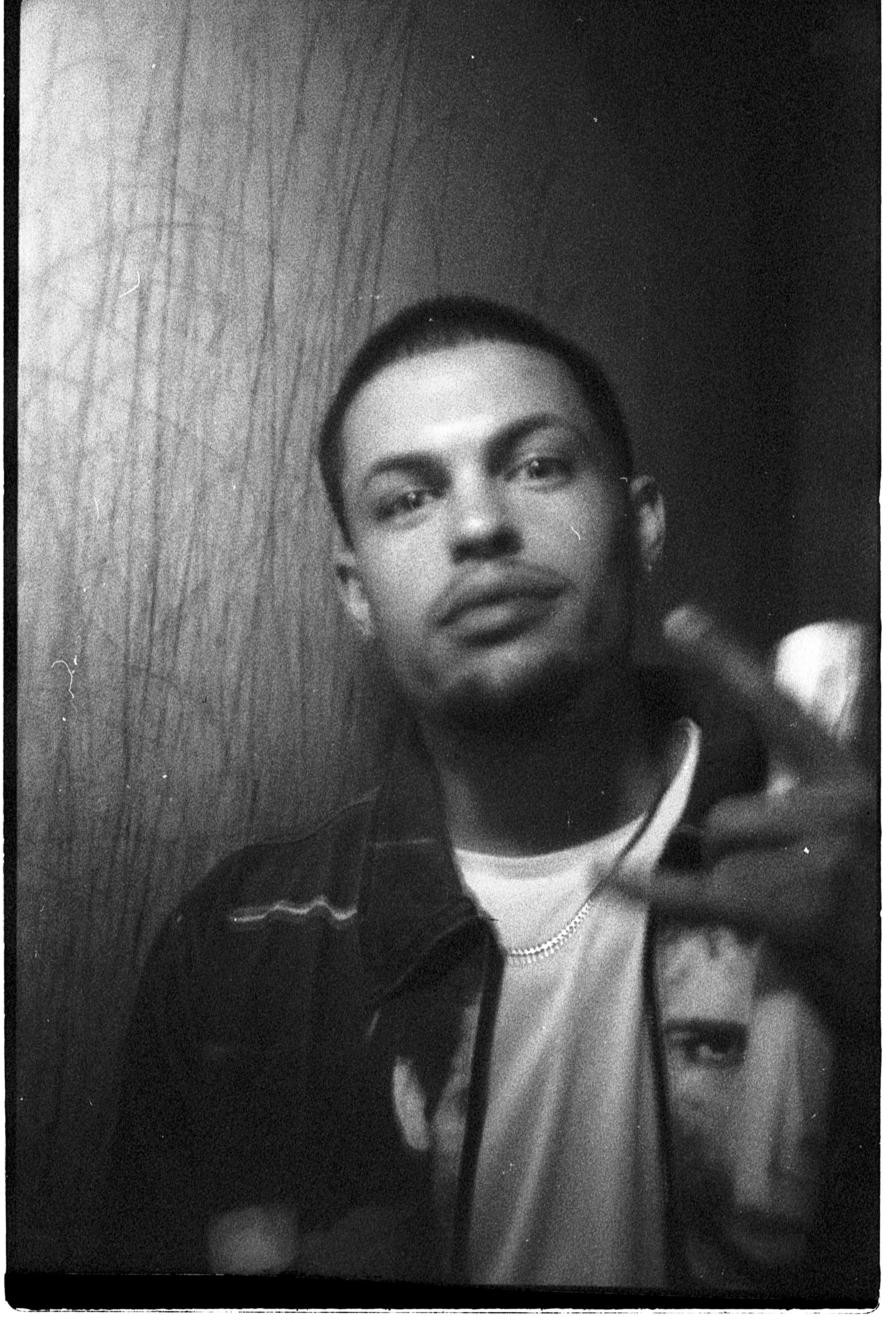 Rapper Doov in a black-and-white press photo from a photobooth, wearing a chain and a graphic jacket, hand raised toward the camera.
