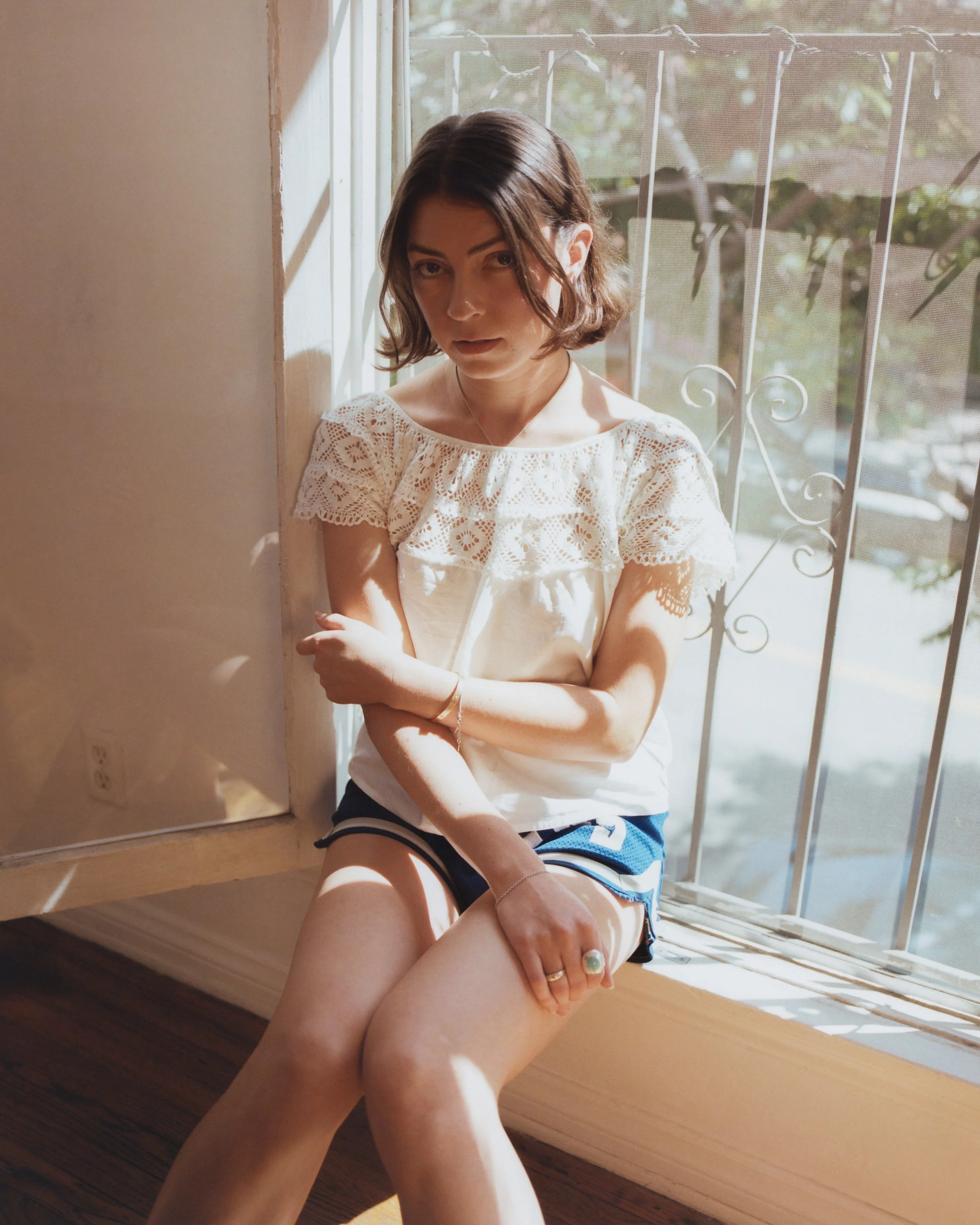 Anna Shoemaker, a white artist with a short brown bob, sits in a sunlit window wearing a white crochet top