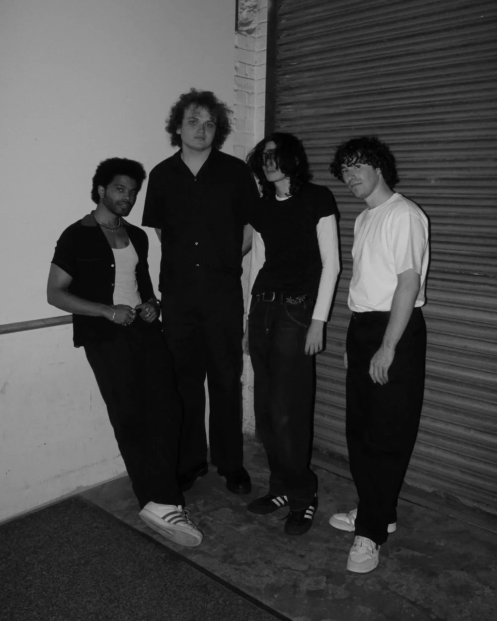 Nautics, a four-piece band of young men in dark outfits, photographed in black and white by an industrial roll-up door at night