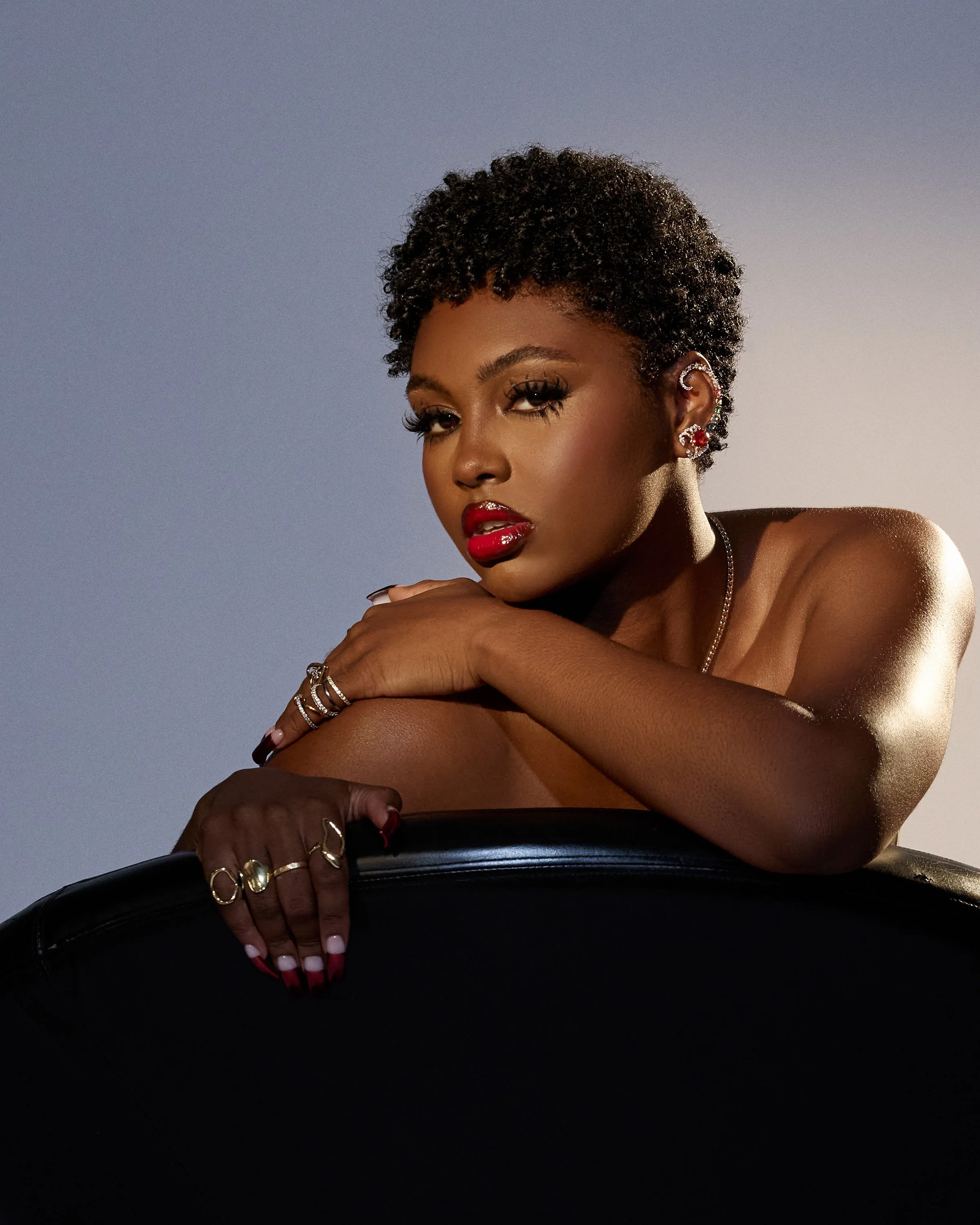 Tiana Major9, a Black artist with short natural curls and a bold red lip, leans over a black leather chair