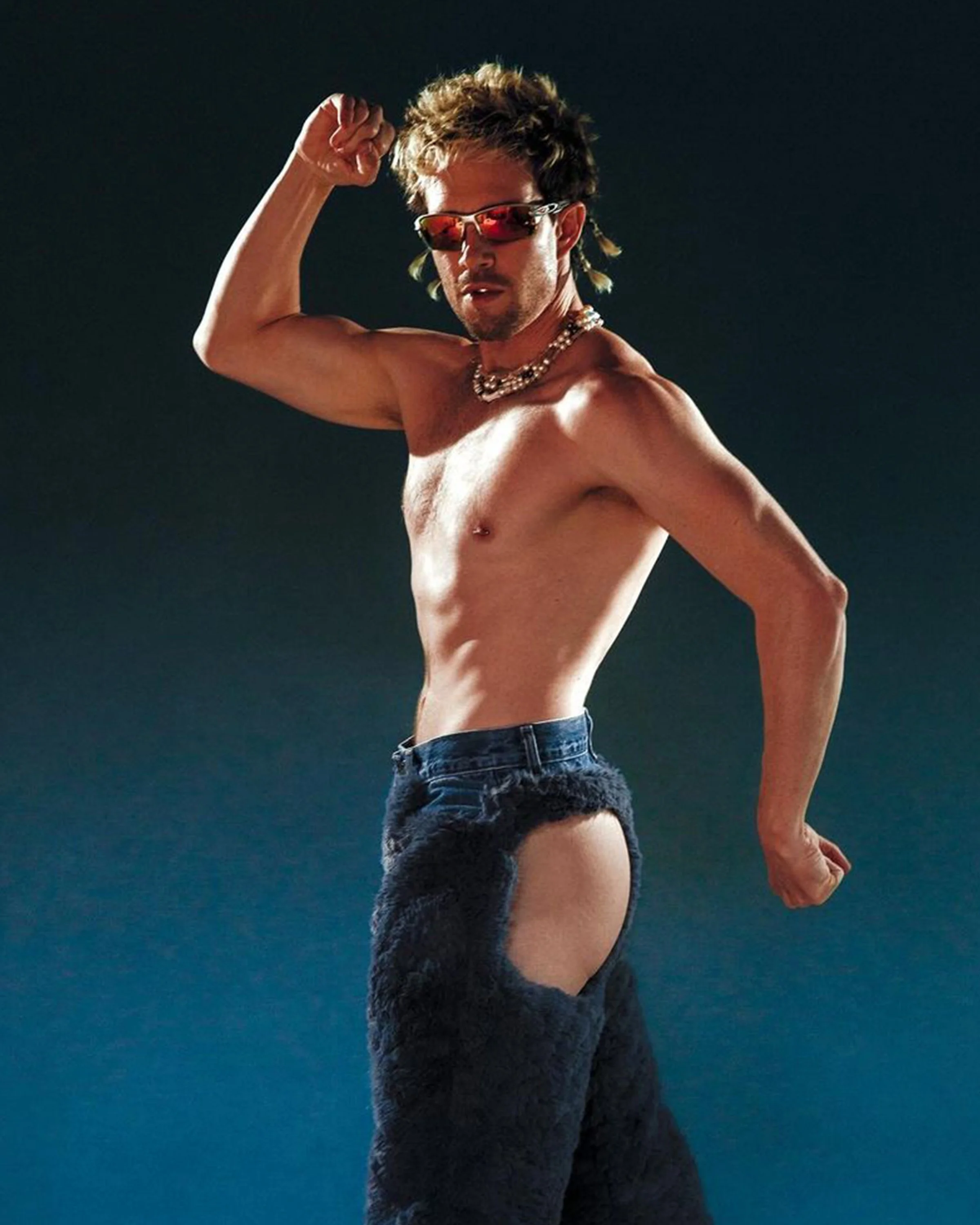 Geordie Kieffer, a white artist, flexes shirtless in red sunglasses, pearls and denim chaps on a blue backdrop