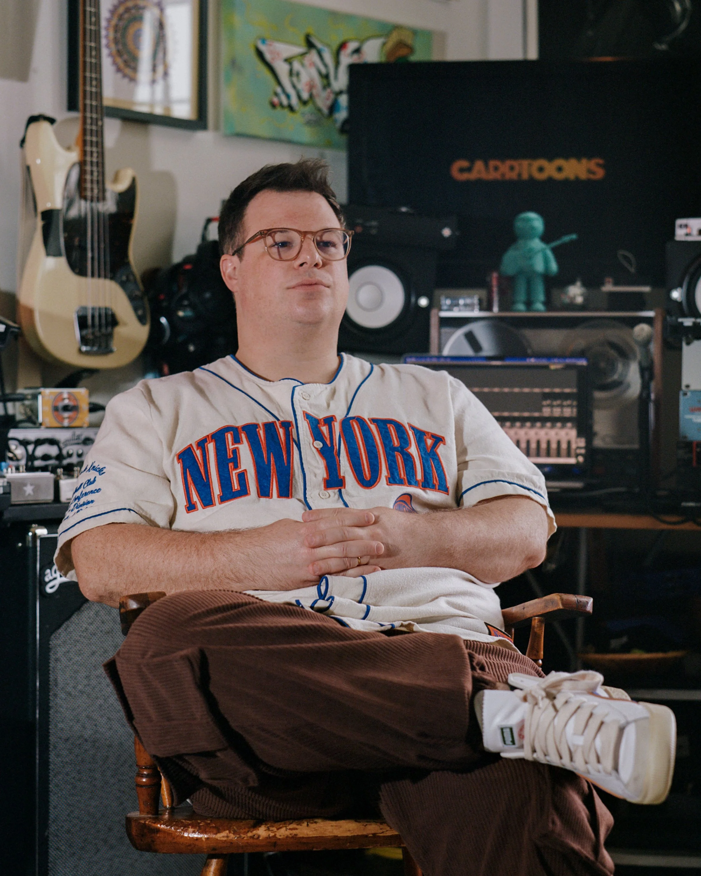 CARRTOONS, a white artist in a New York baseball jersey, sits in his studio beside a bass guitar and monitors