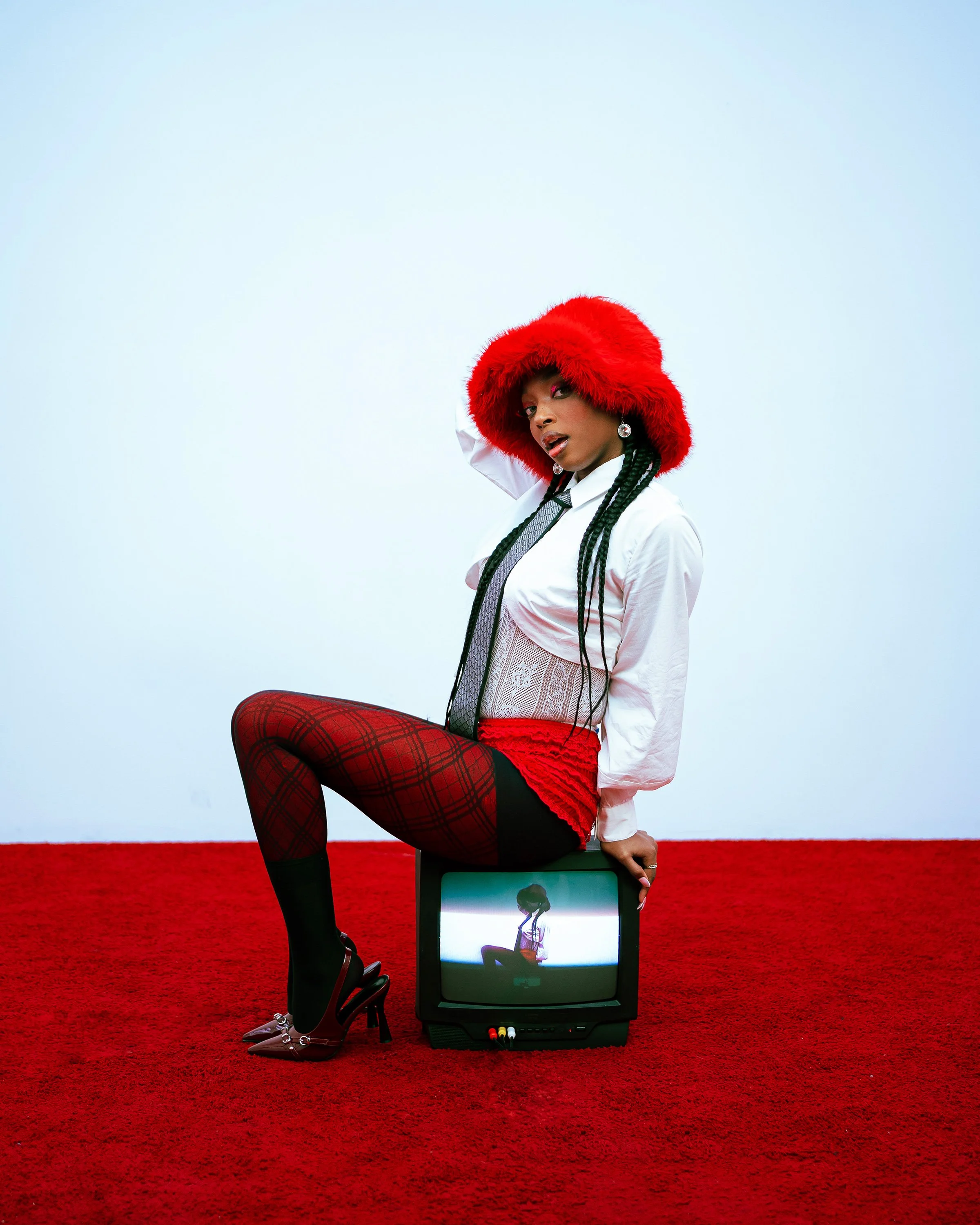 Shelailai, a Black artist in a red fur hat and plaid tights, perches on a vintage TV showing her own image