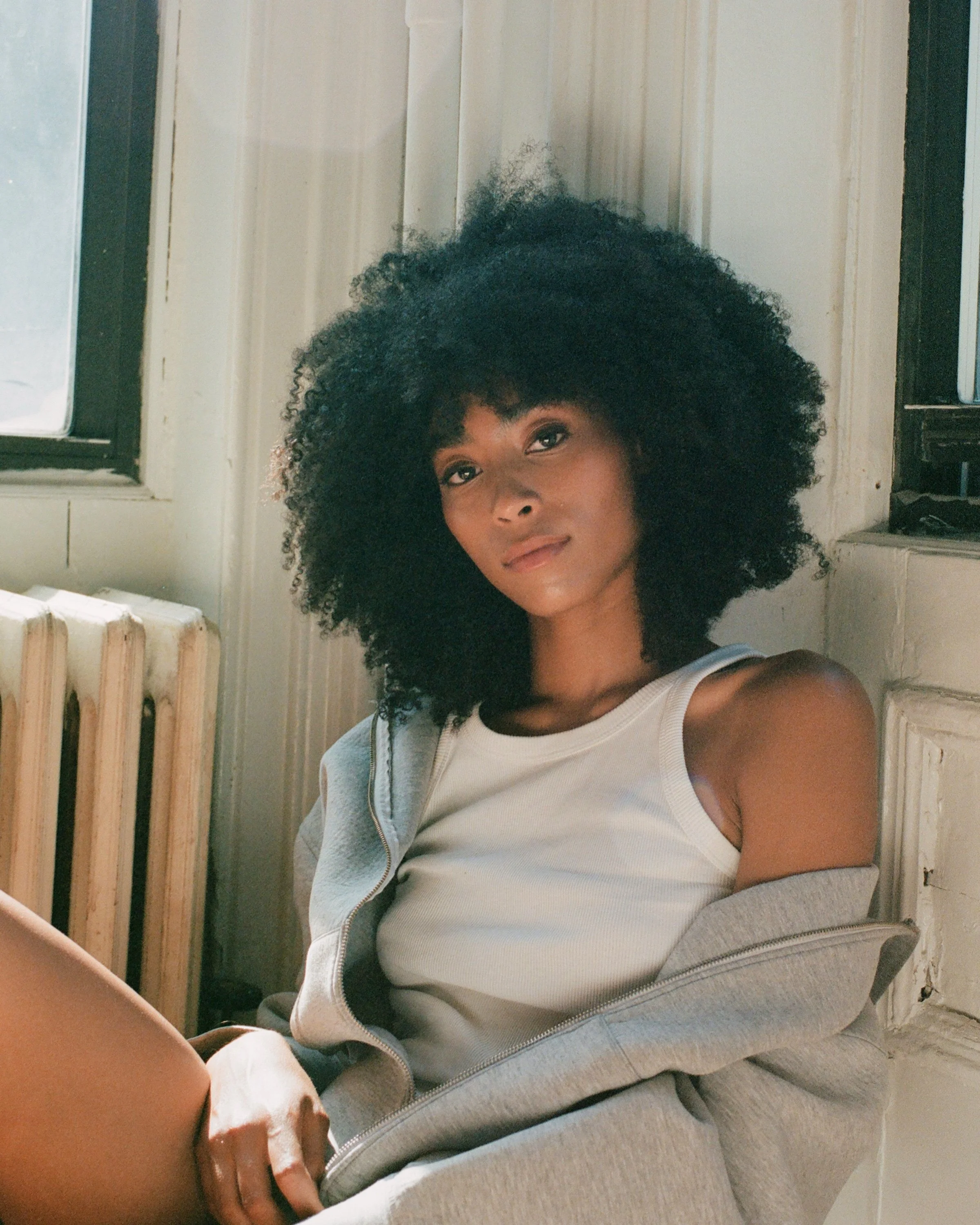 Phoenix James, a Black artist with a natural afro, sits by a sunlit window in a gray tank and hoodie