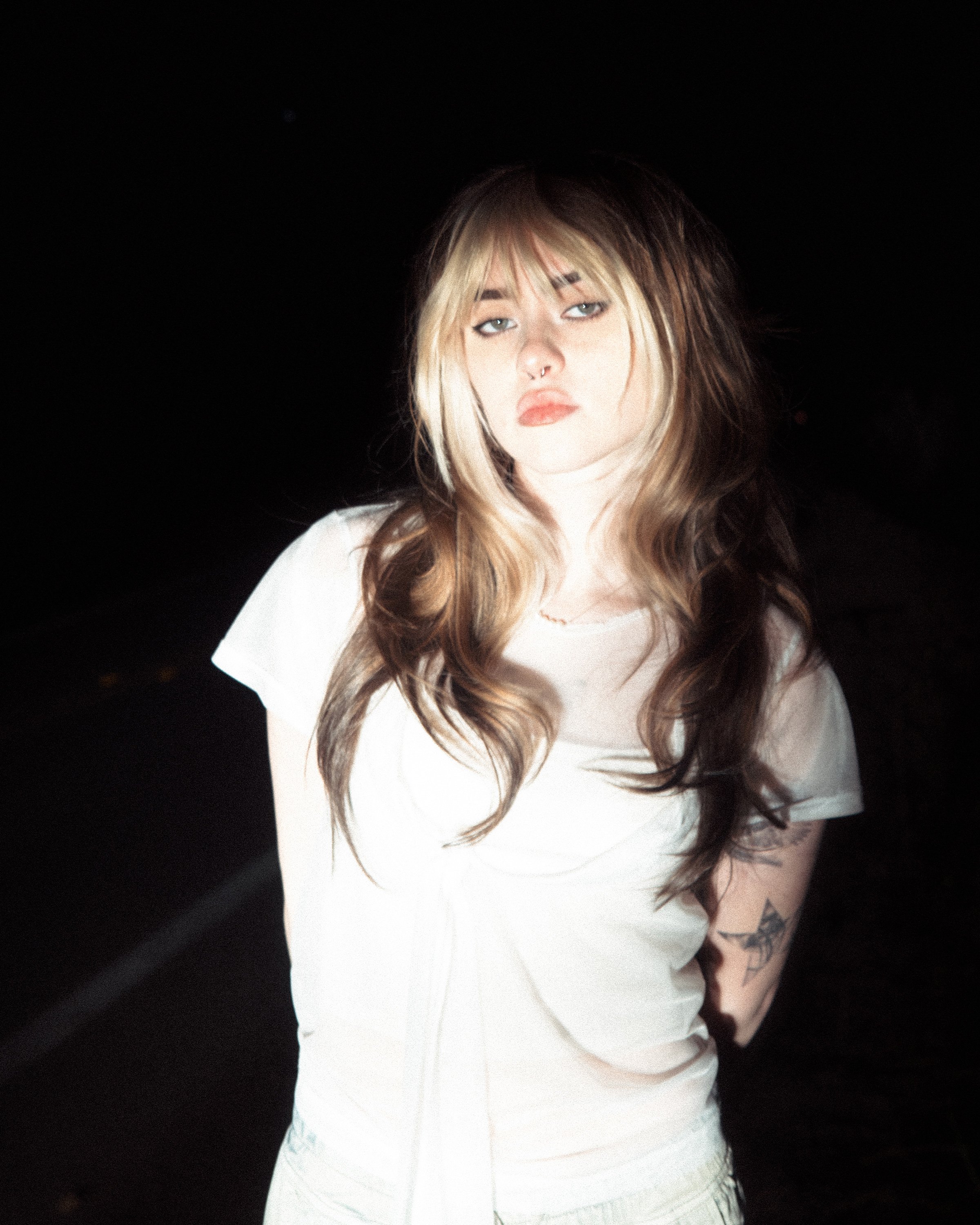 Ella Boh, a white blonde artist, photographed at night in a white tee, flash catching a forearm tattoo