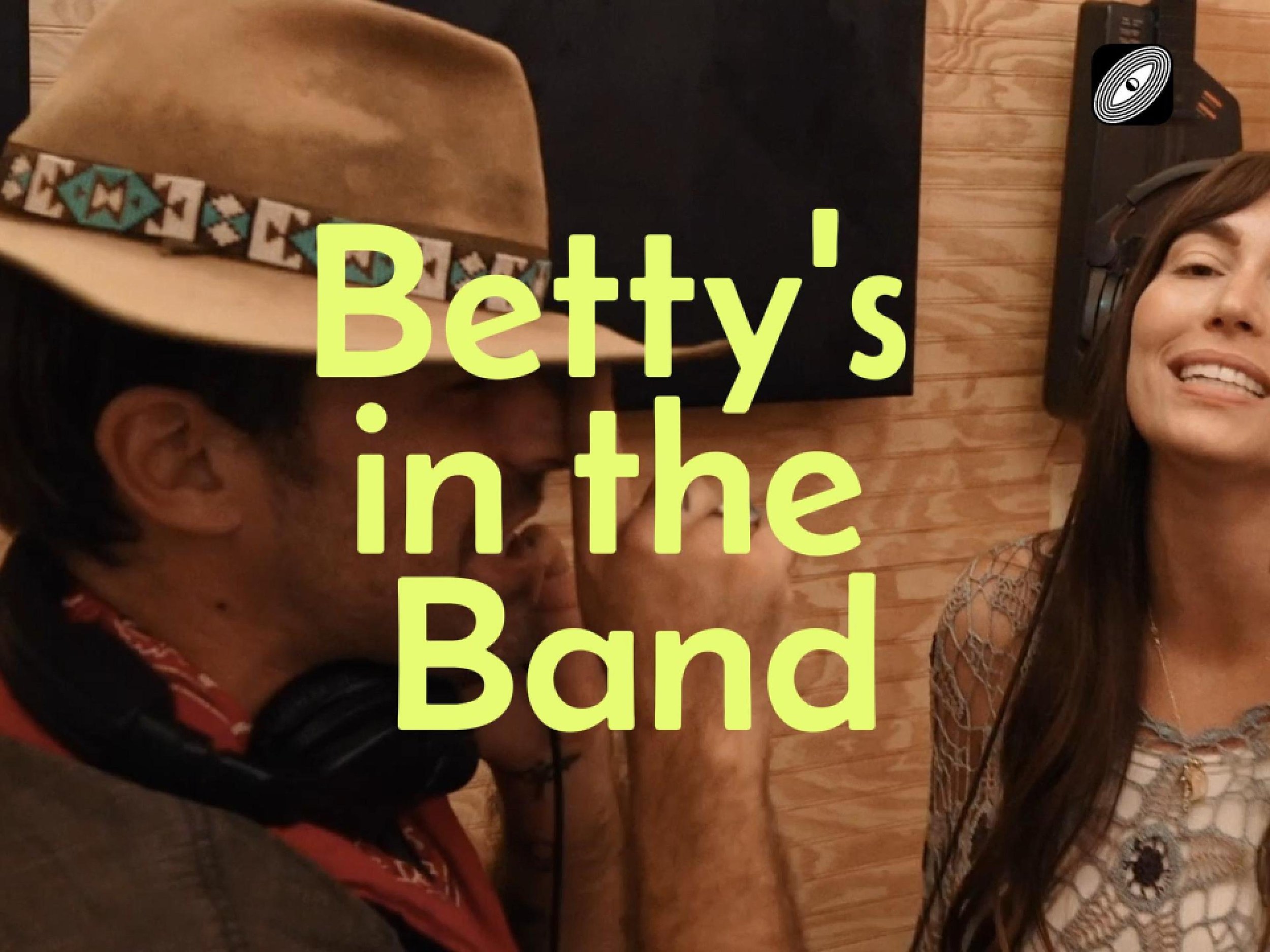A woman wearing headphones and a woman wearing a cowboy hat with a turquoise and white band, smiling. The background is a wooden paneled wall with a black object in the top right corner.