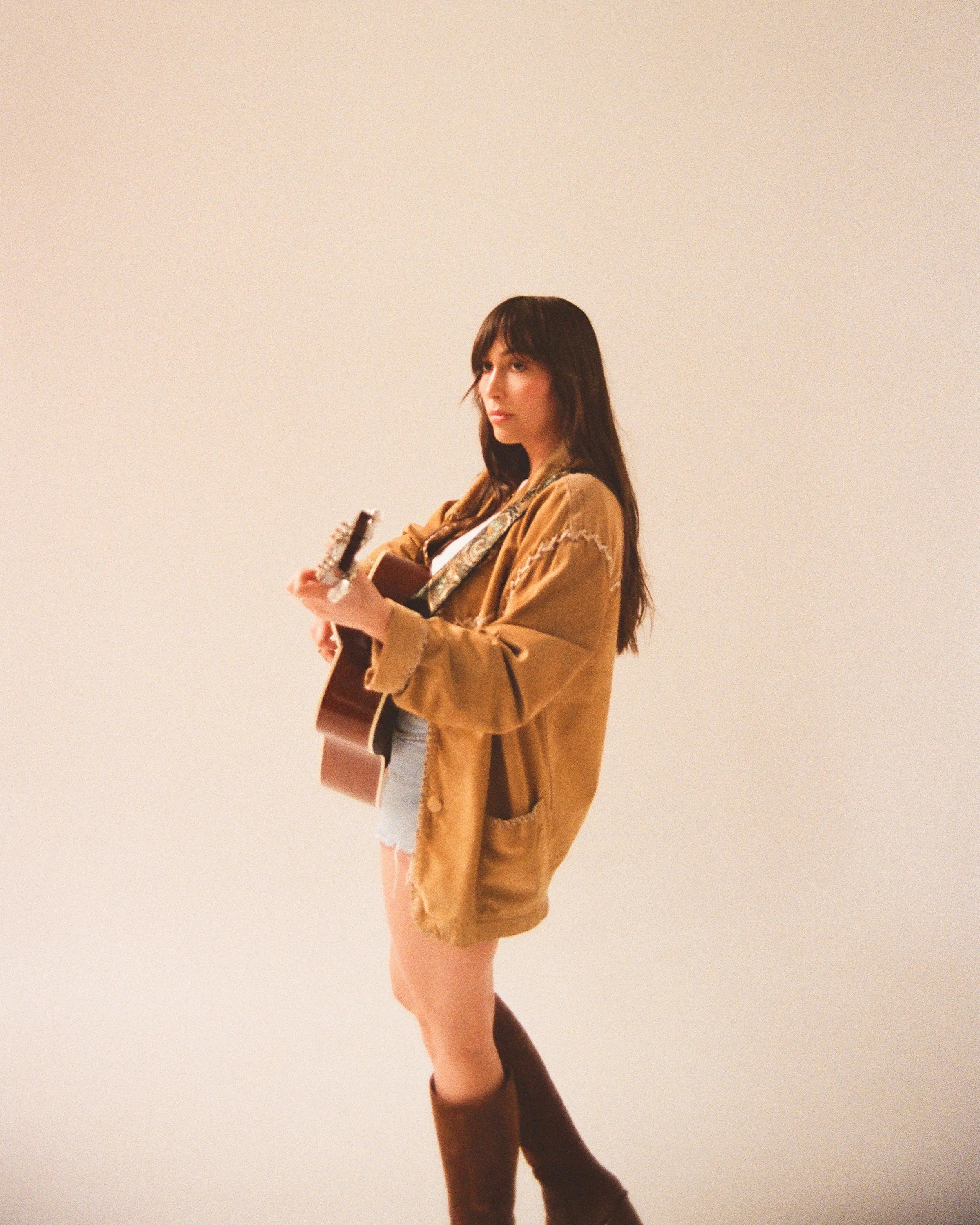 Abigail Rose, a fair-skinned artist with dark hair and bangs, holds an acoustic guitar in a camel cardigan and boots