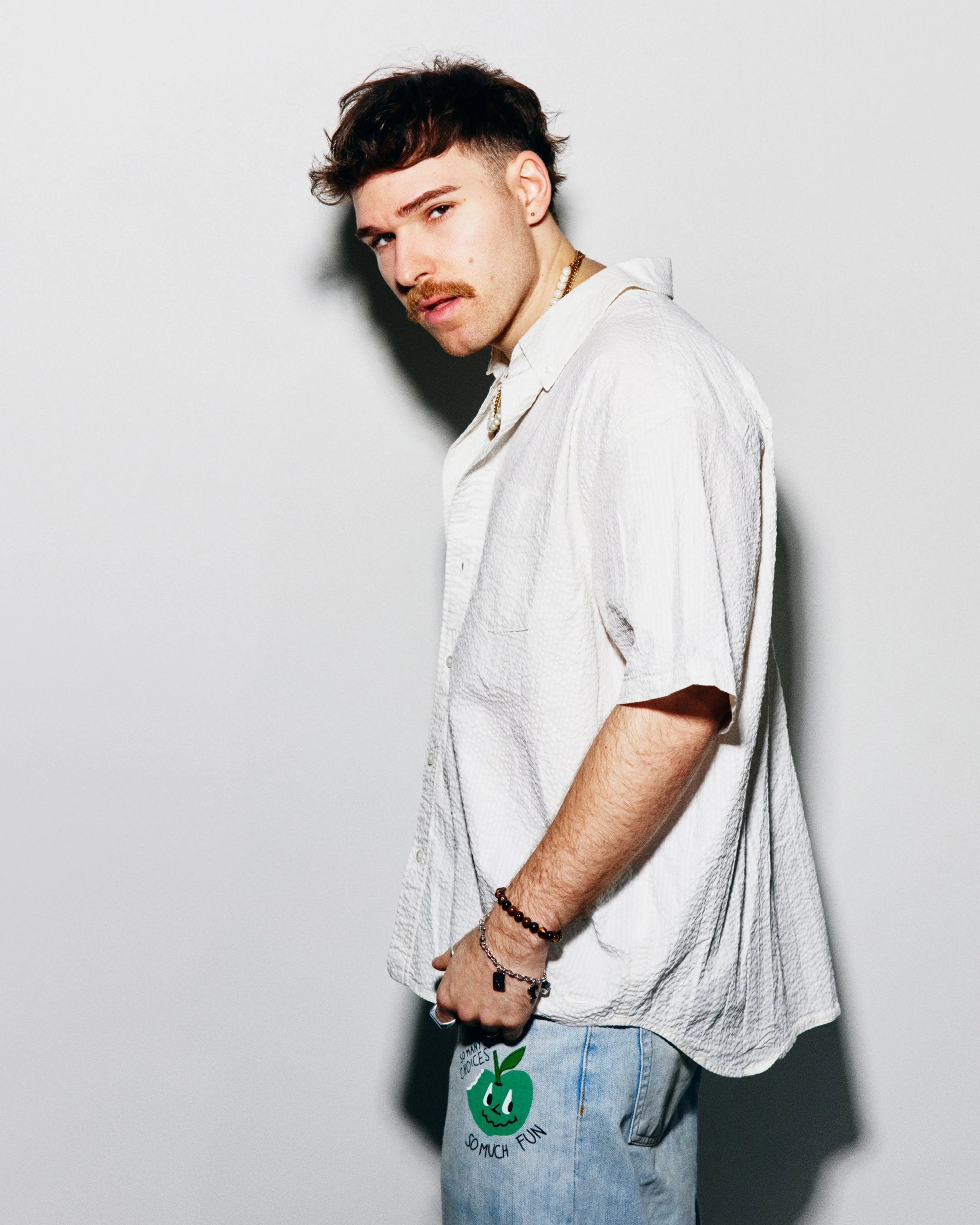 Ely Oaks, a white artist with a mustache, stands in profile in an oversized cream shirt and graphic-print jeans