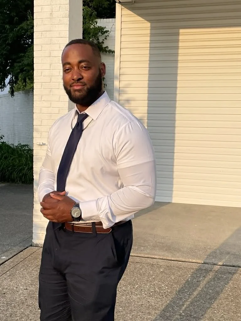 A well-dressed man with a beard and short hair, wearing a white dress shirt, dark tie, black pants, and a watch, stands outdoors during sunset next to a white brick wall and a rolled-up garage door.