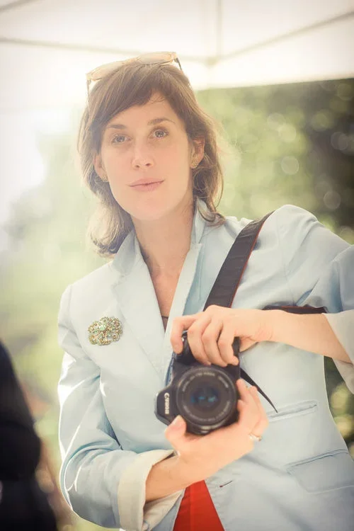 Woman with short brown hair wearing a light blue blazer and sunglasses on her head, holding a camera and taking a selfie outdoors.