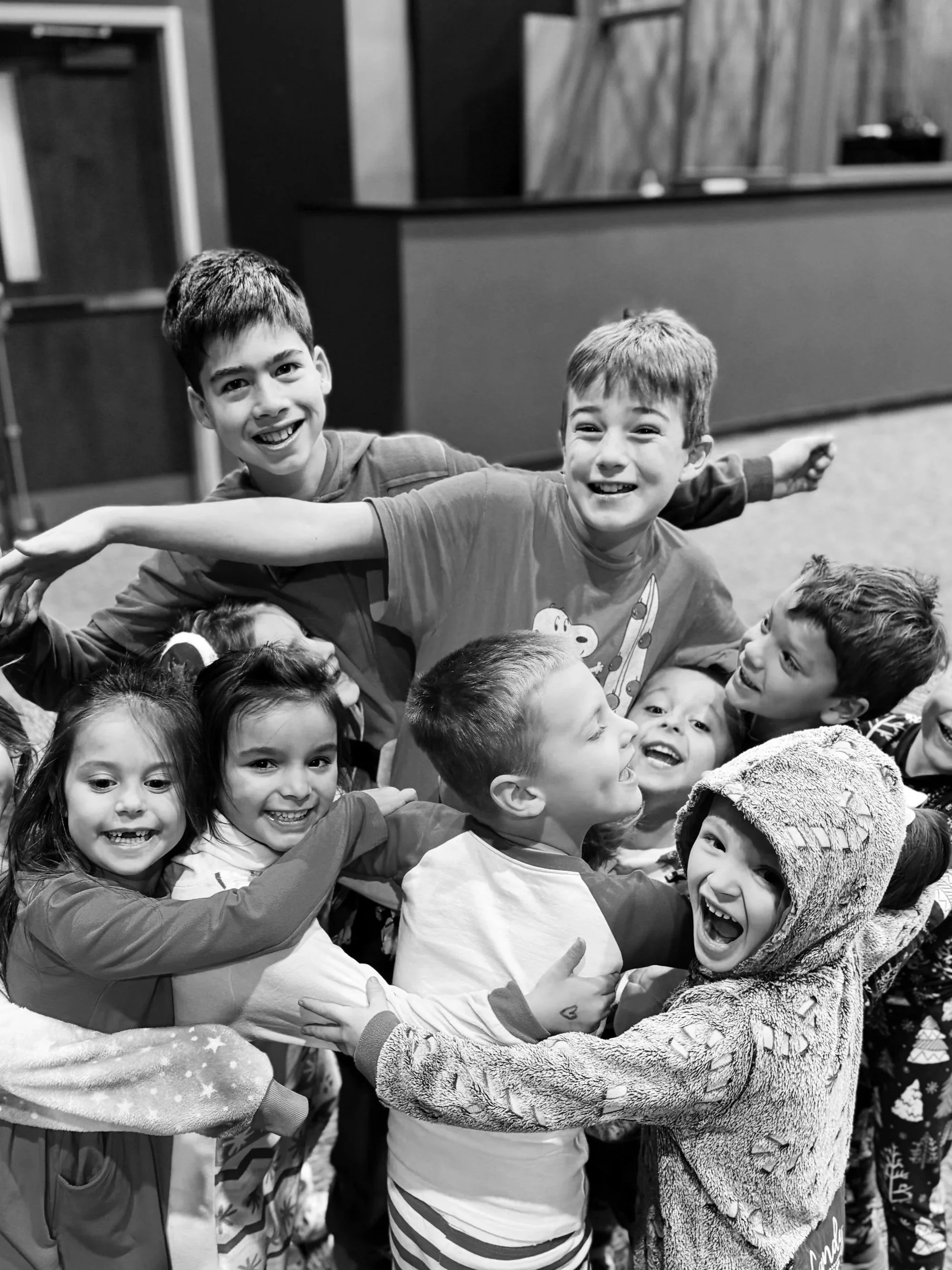 A group of children hugging and playing together, smiling and laughing, indoors.