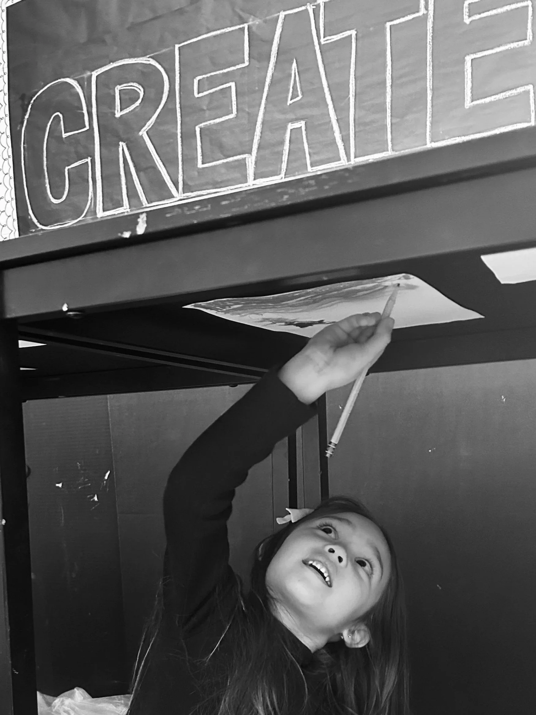 A young girl is reaching up with a pencil to the underside of a table with a sign that reads 'CREATE' in large letters. She is looking up at the sign with a joyful expression.