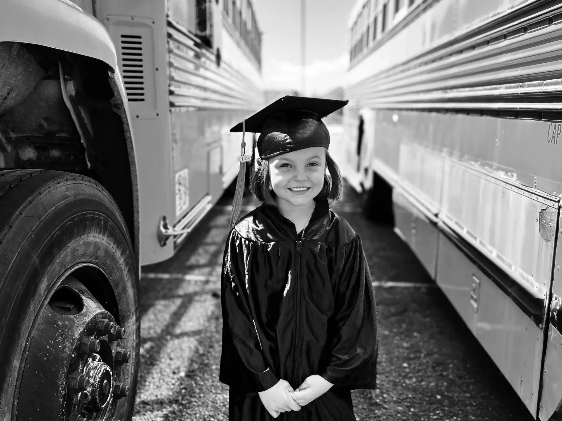 A young girl in a graduation gown and mortarboard, standing between two large buses on a sunny day, smiling at the camera.