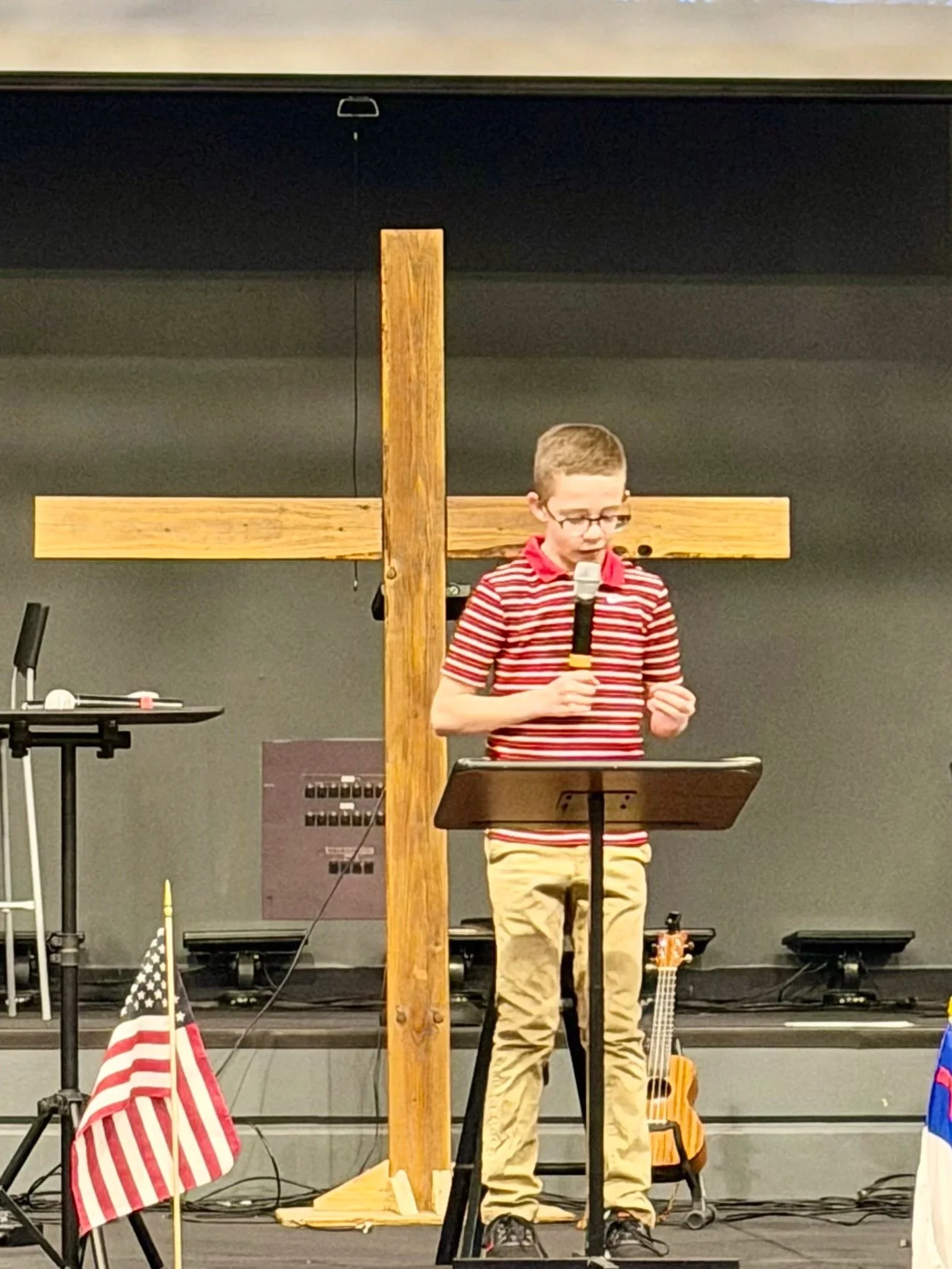 A boy with glasses, wearing a red and white striped polo shirt and khaki pants, standing at a music stand, holding a microphone and speaking in front of a large wooden cross, with American flags and a guitar nearby.
