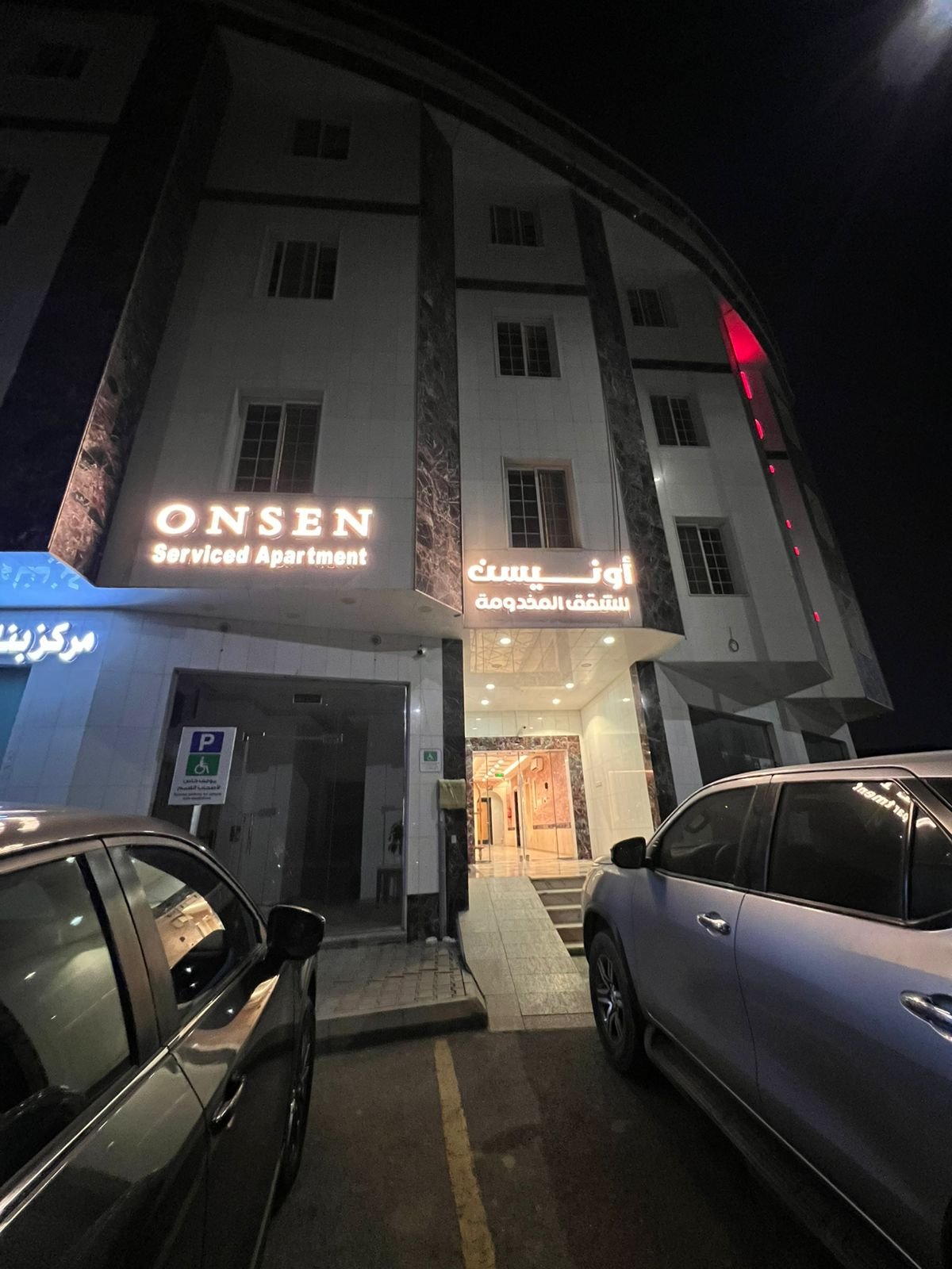 Night view of a multi-story apartment building with illuminated signs in English and Arabic, indicating 'ONSEN Serviced Apartment,' with parked cars in the foreground and a parking sign.