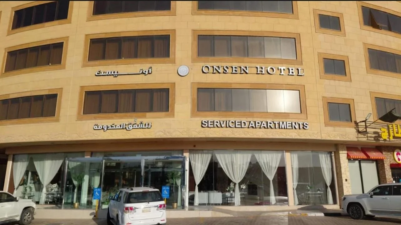 The exterior of Onsen Hotel with signage in English and Arabic for serviced apartments, a glass entrance, and parked cars in front.