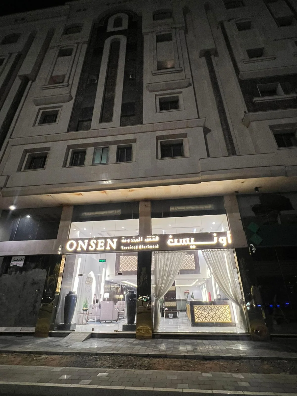 Nighttime view of the entrance to a hotel named 'ONSEN' with a sign indicating it is a serviced apartment. The hotel has large glass windows with curtains inside, and a cozy lobby visible with pink chairs, decorative vases, and warm lighting.