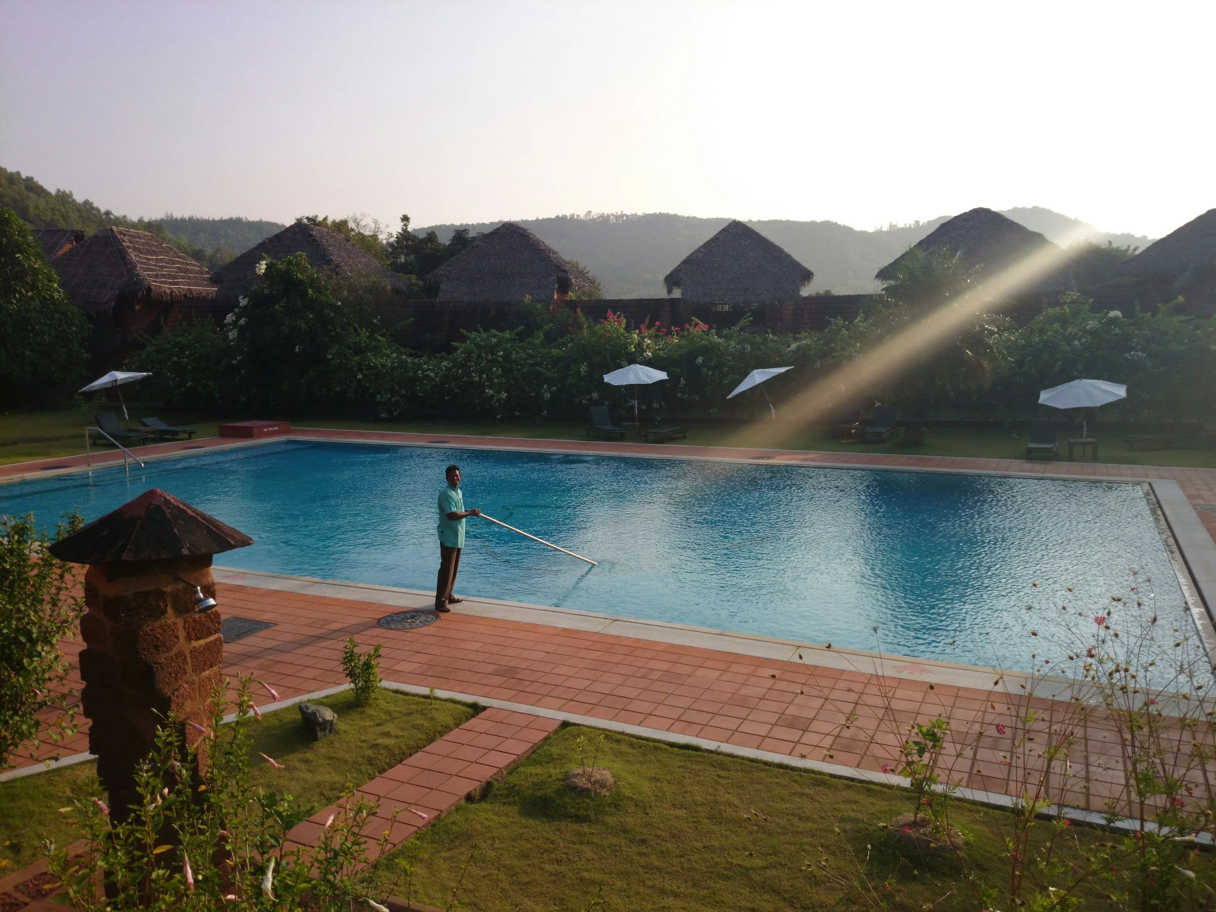 Man cleaning a swimming pool in a resort with thatched-roof cottages and greenery in the background during sunset.