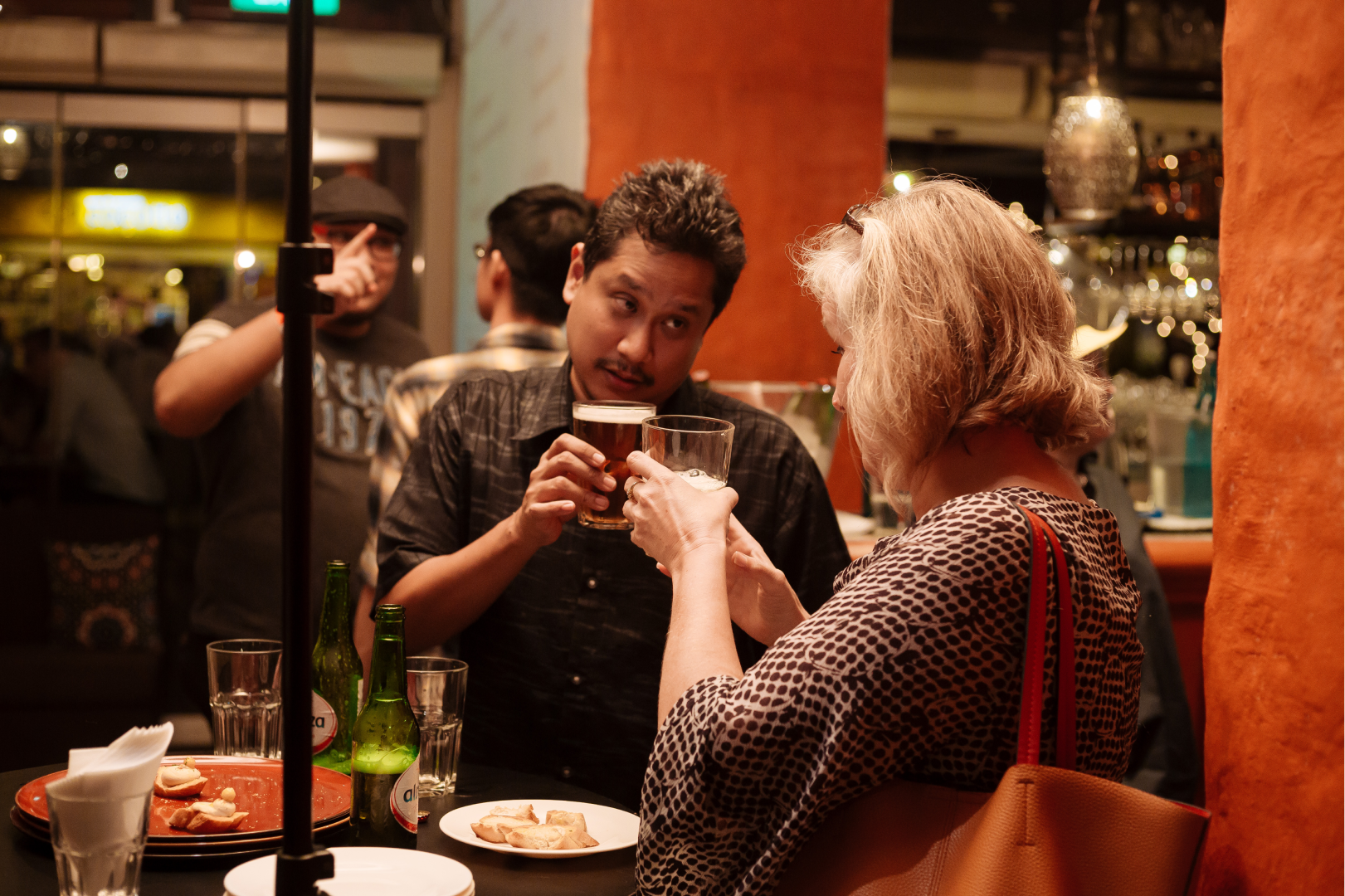 A man and a woman talking and holding drinks at a restaurant or bar, with other people and bar setup in the background.