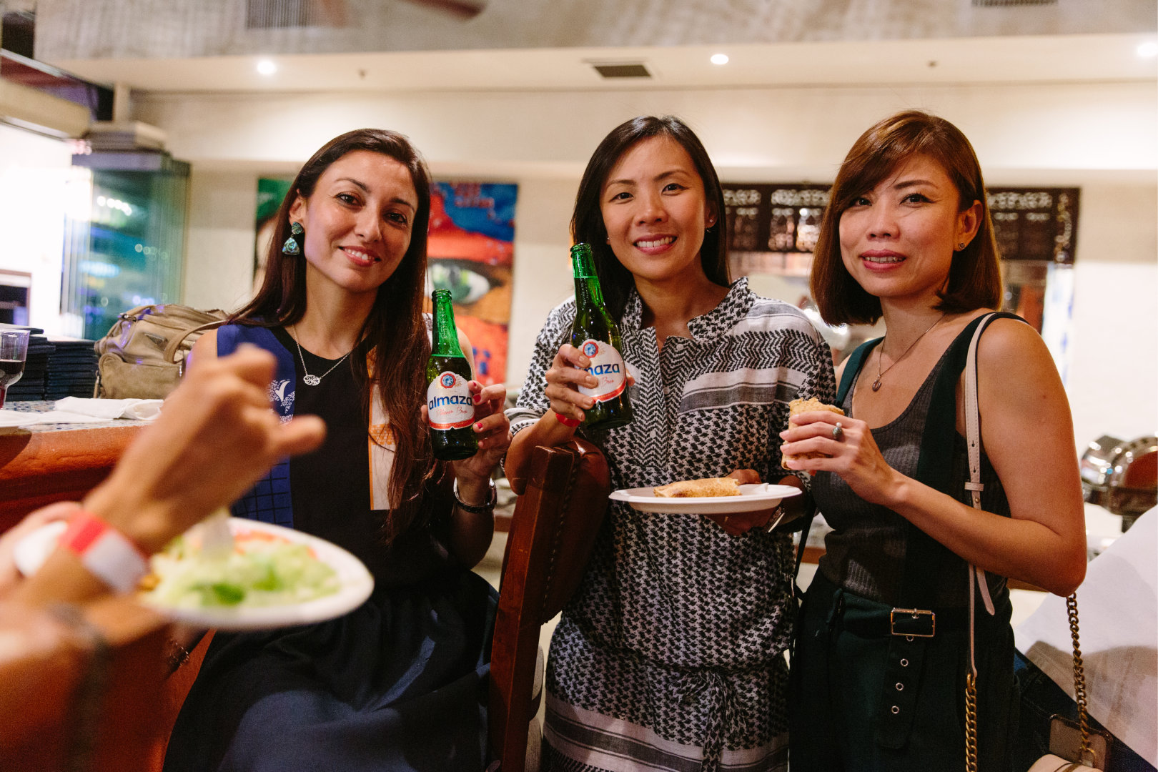 Three women smiling at a social gathering, holding drinks and food, in a restaurant or bar setting.