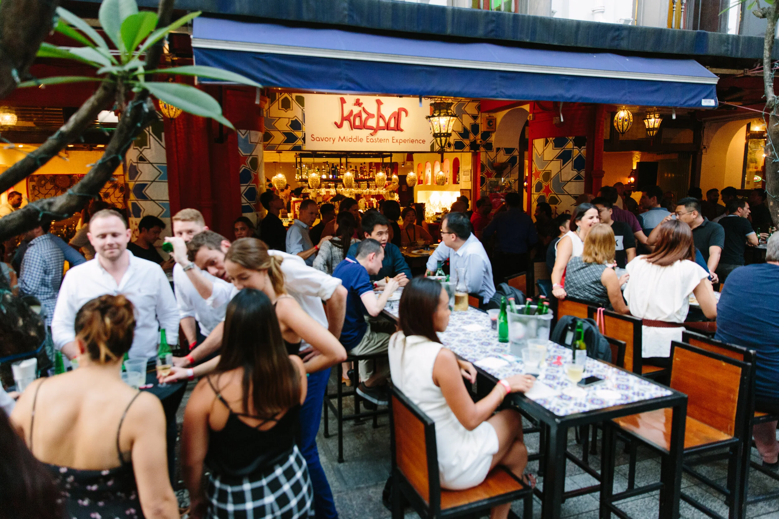 Crowded restaurant with many people dining and socializing, with colorful Middle Eastern decor and a bar in the background.