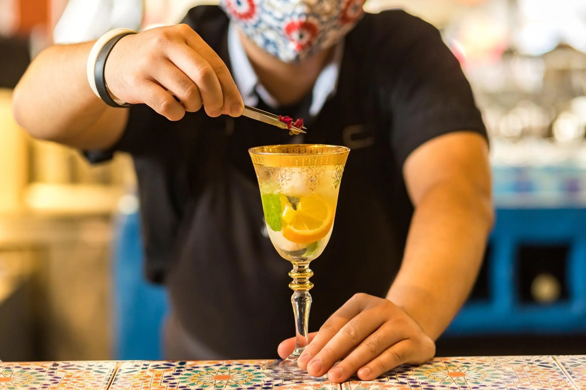 A bartender wearing a floral face mask decorates a colorful cocktail with a small flower using tweezers.