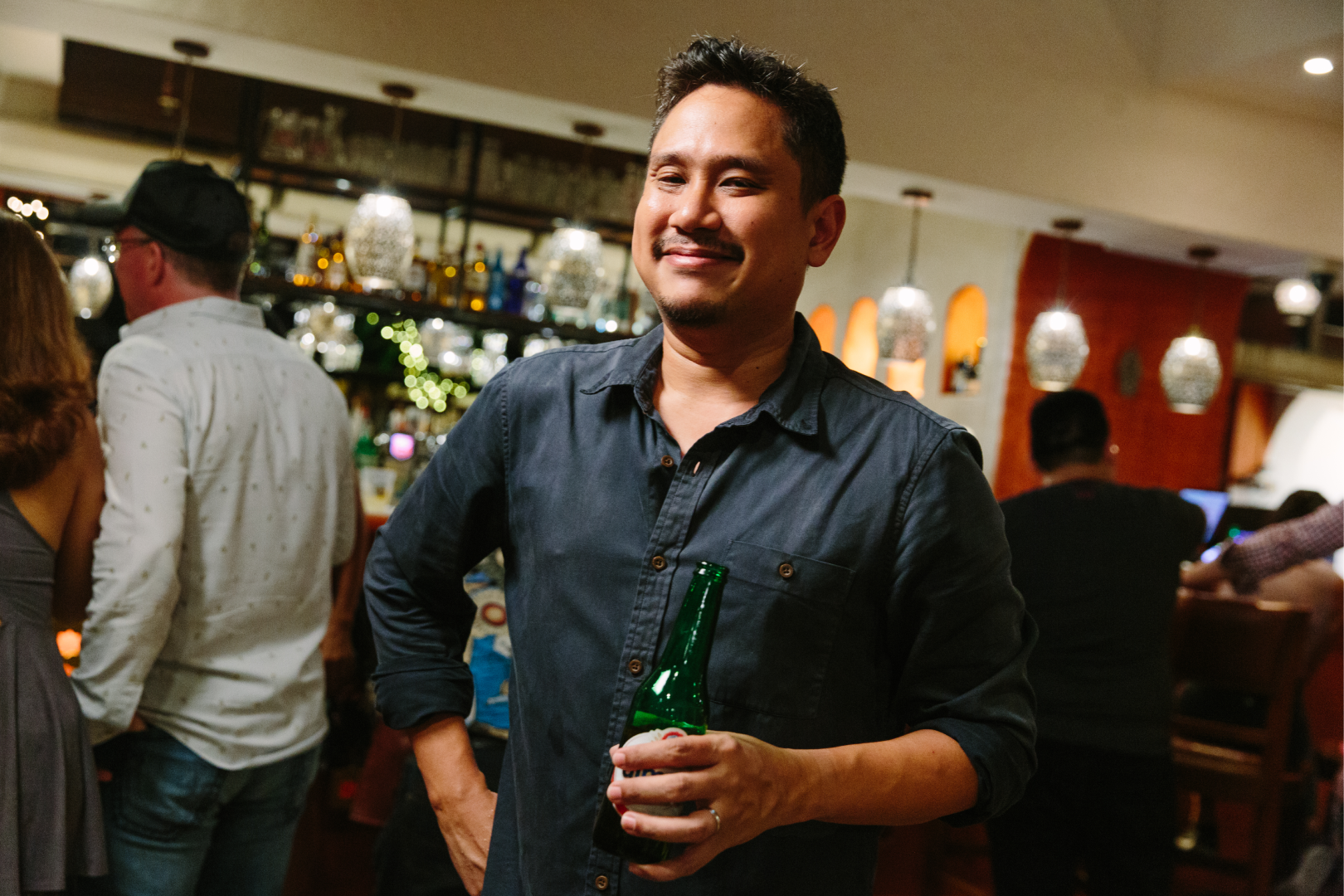 A man smiling holding a beer bottle at a lively bar or pub with people in the background.