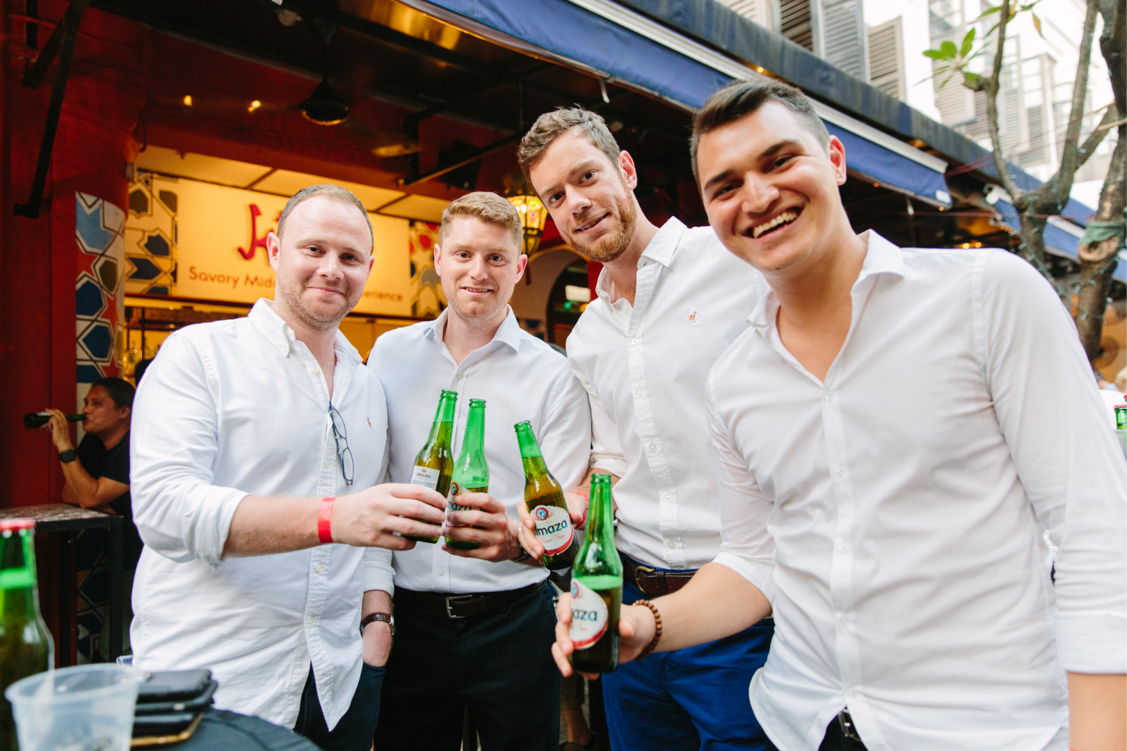 Four young men in white shirts smiling and holding beer bottles at an outdoor event or bar.
