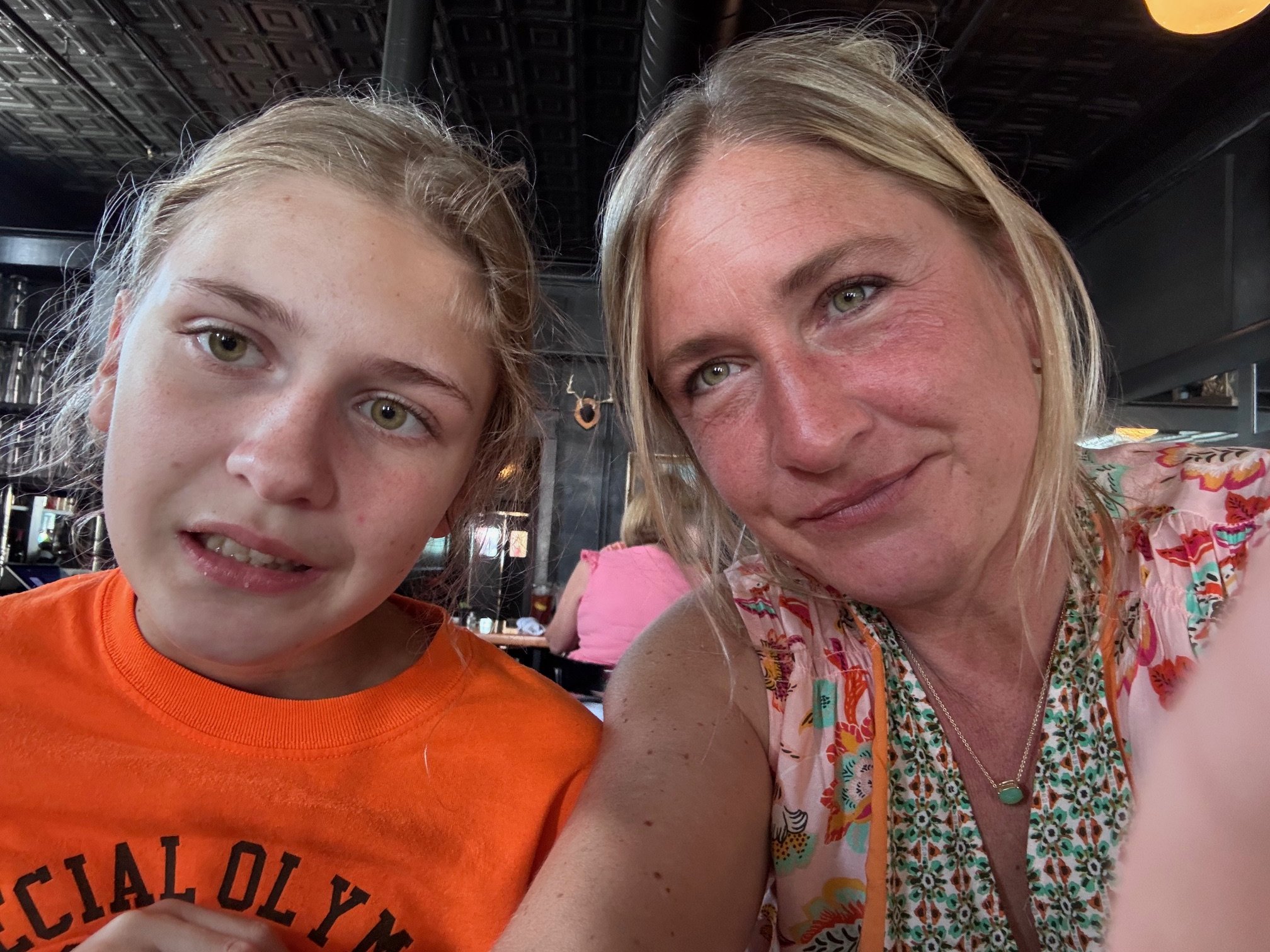 A woman and a young girl taking a close-up selfie in a restaurant or cafe. The woman has blonde hair, blue eyes, and is wearing a floral top with a necklace. The girl has blonde hair, greenish eyes, and is wearing an orange sleeveless top.