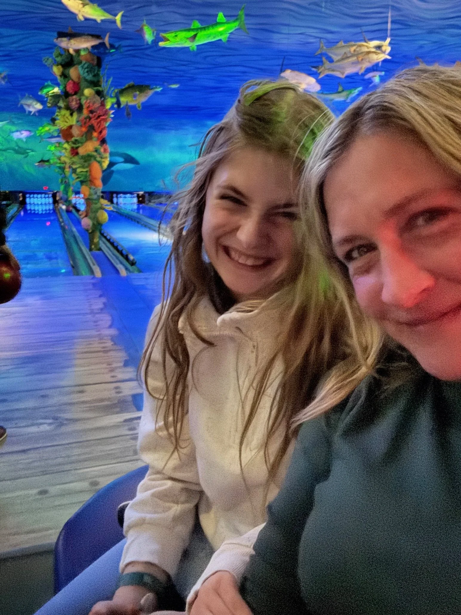 Two women taking a selfie inside an aquarium with a large tank filled with colorful fish and coral in the background.