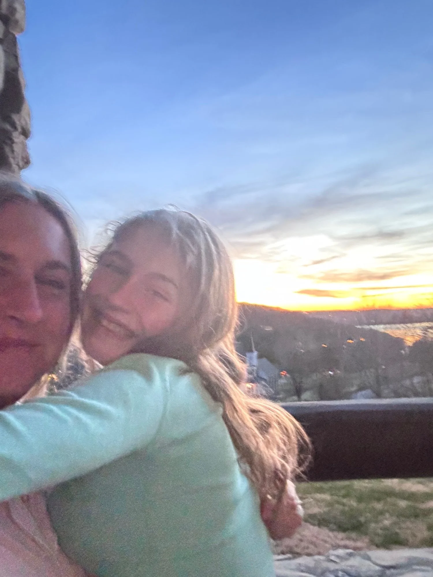 Two women smiling and hugging outdoors during sunset, with a scenic landscape and houses in the background.