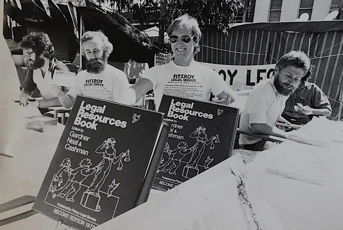 Four people promoting Fitzroy Legal Service with signs 'Legal Resources Book' in front,  two are looking towards the camera; one person is wearing sunglasses.