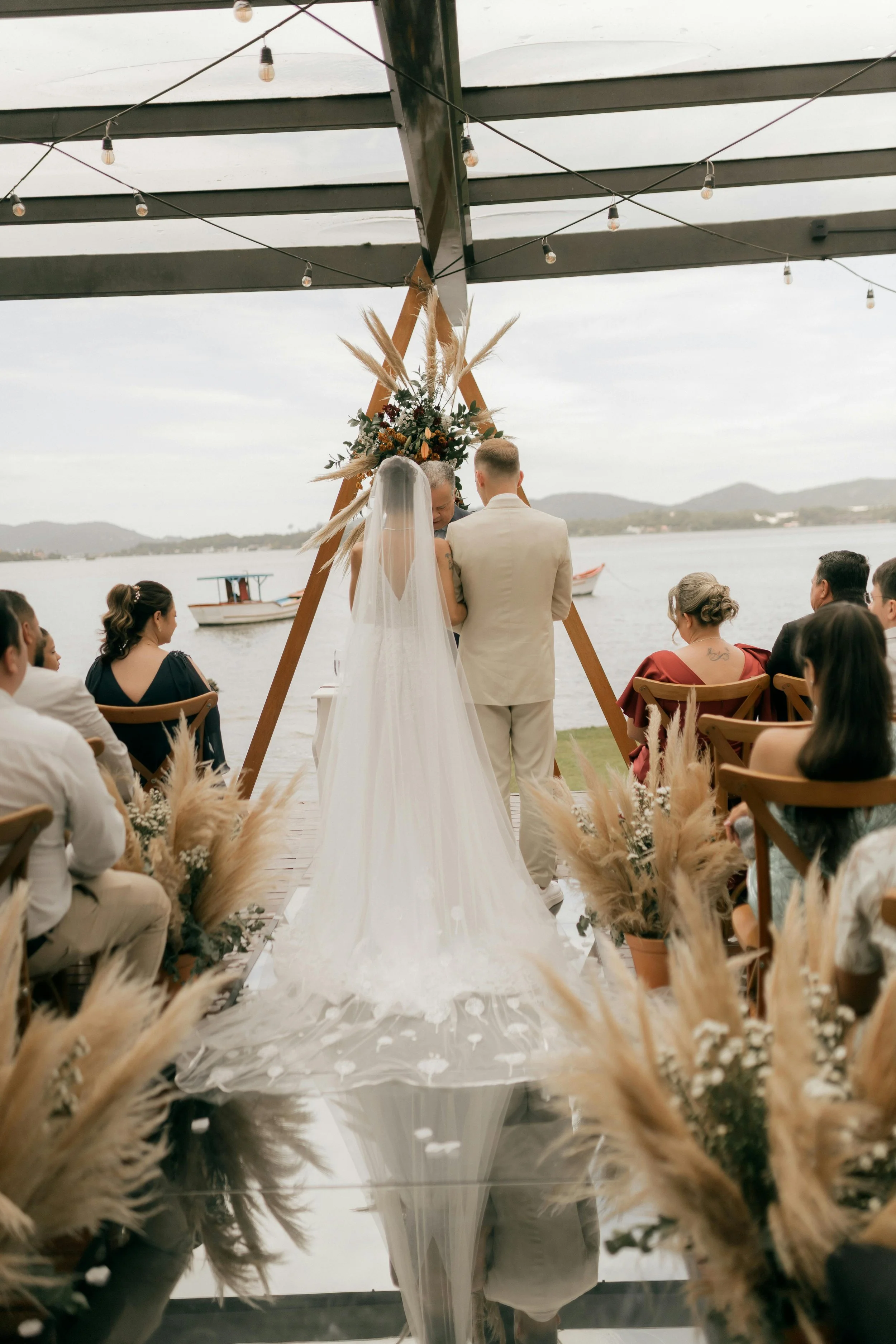 A wedding ceremony taking place outdoors on a lakeside with a couple standing under a floral arch, surrounded by seated guests, and boats on the water in the background.