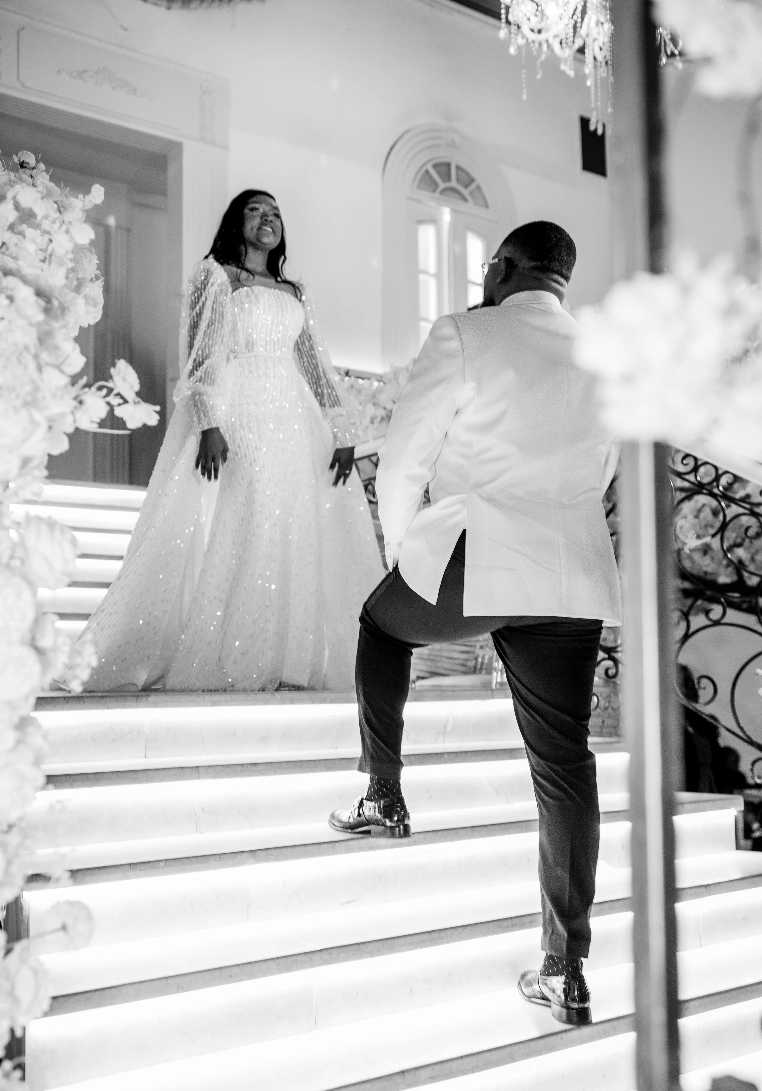 Black and white photo of a man proposing to a woman on a grand staircase decorated for a wedding or formal event, with a chandelier above.