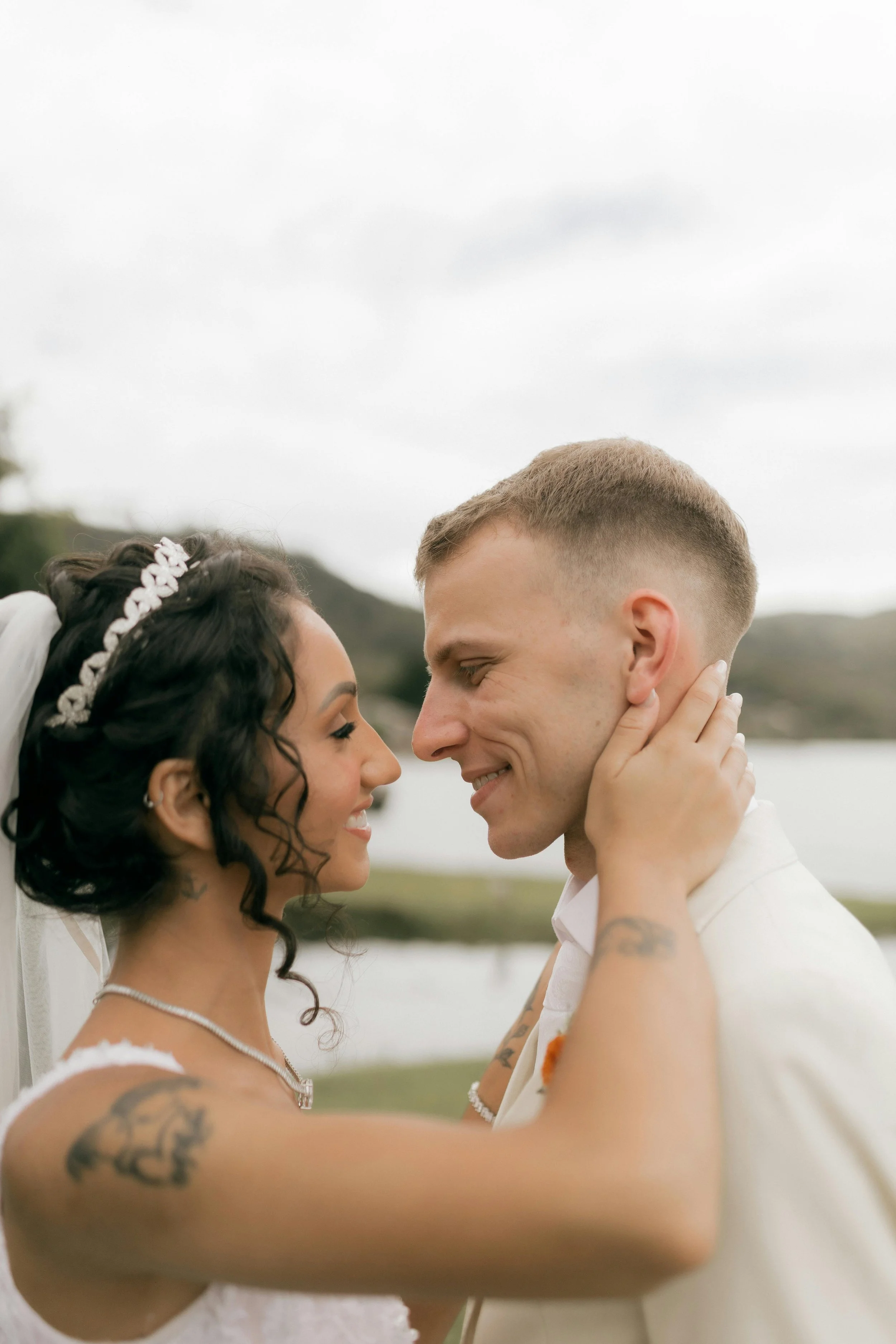 A bride and groom are close together, smiling and looking into each other's eyes outdoors on a cloudy day.