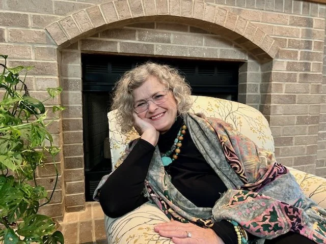 A smiling woman with curly gray hair and glasses, sitting comfortably in a patterned armchair in front of a brick fireplace, with a potted plant nearby.