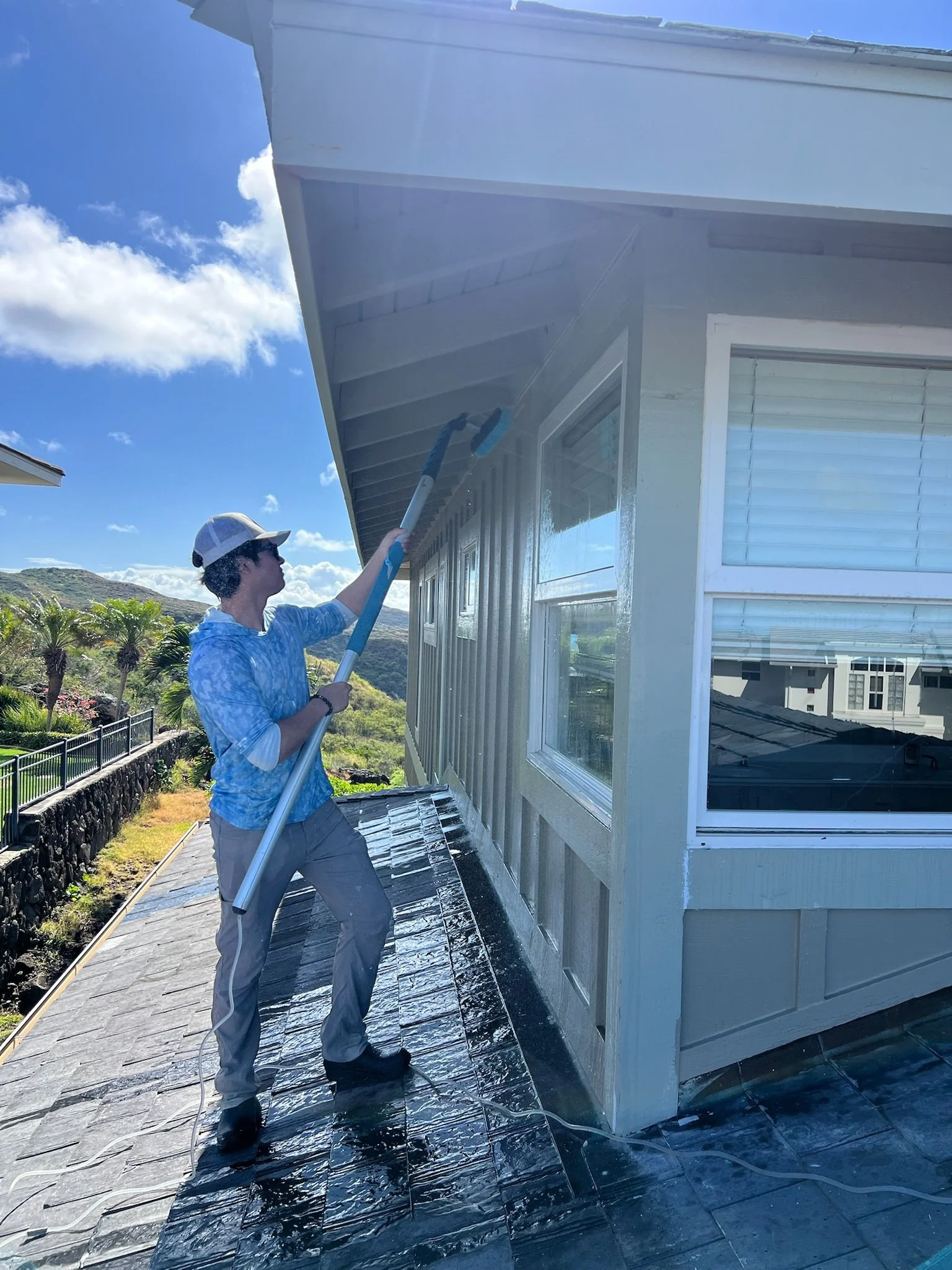 Person cleaning or washing a house's exterior wall with a pressure washer or hose, on a sunny day.