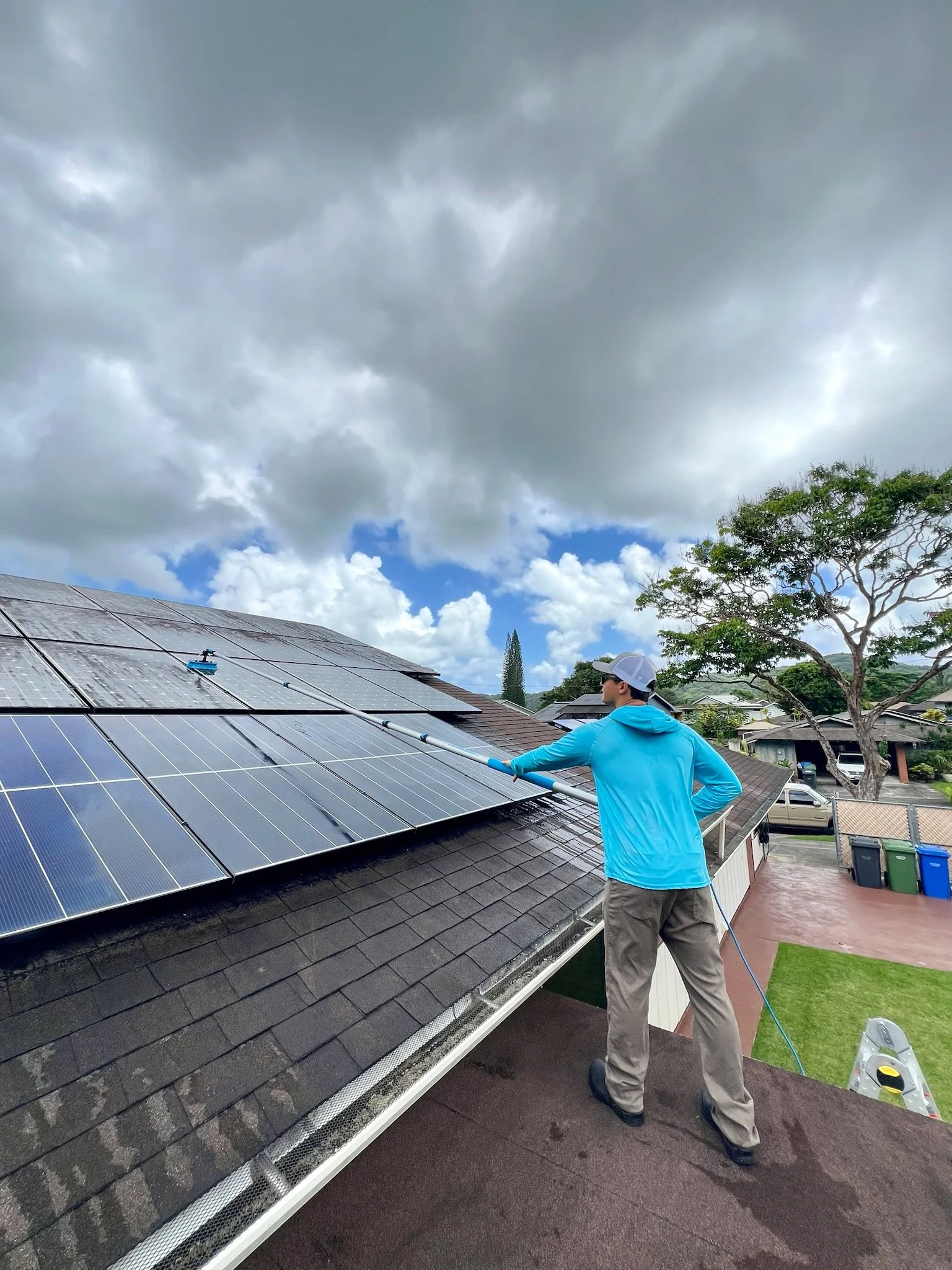 A person in a blue hoodie, gray pants, and a gray cap is cleaning solar panels on a rooftop with a water hose on a cloudy day with dark clouds in the sky and trees and houses in the background.