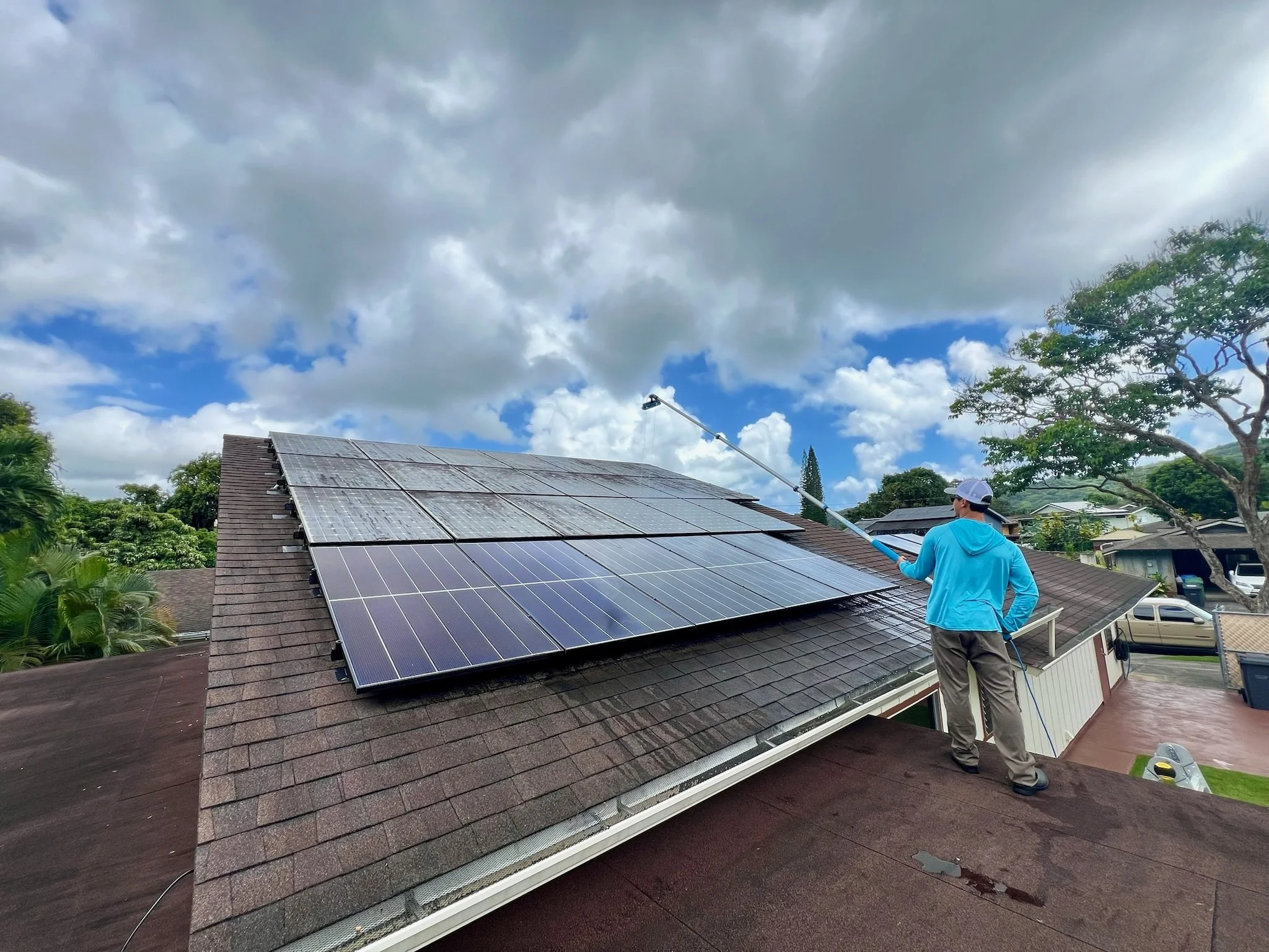 A person in a blue hoodie and cap is cleaning solar panels on a roof with a long-handled cleaning tool. The rooftop has solar panels installed, and the sky is partly cloudy with some blue visible.