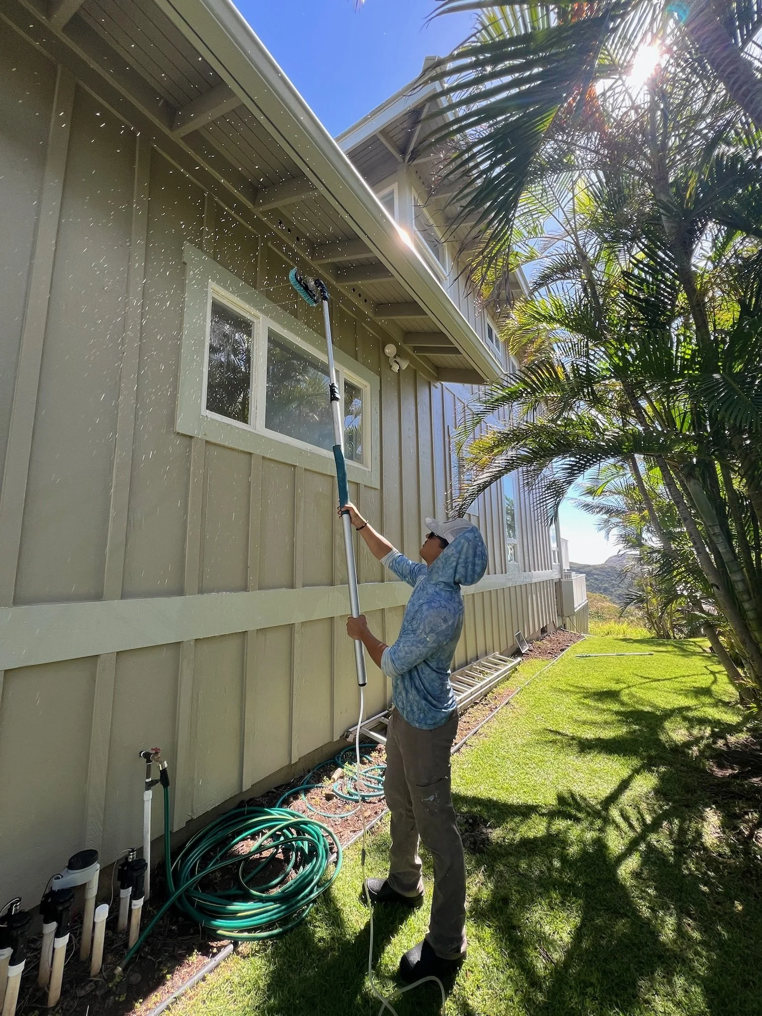 A person cleaning the exterior of a house using a long-handled brush, with water splashing around, in a backyard with palm trees and a grassy lawn.