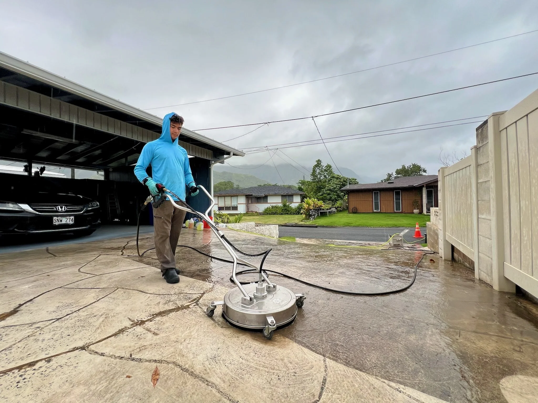 A man wearing a blue hoodie and gloves is operating a floor cleaning machine on a concrete driveway in a residential neighborhood on a cloudy day.