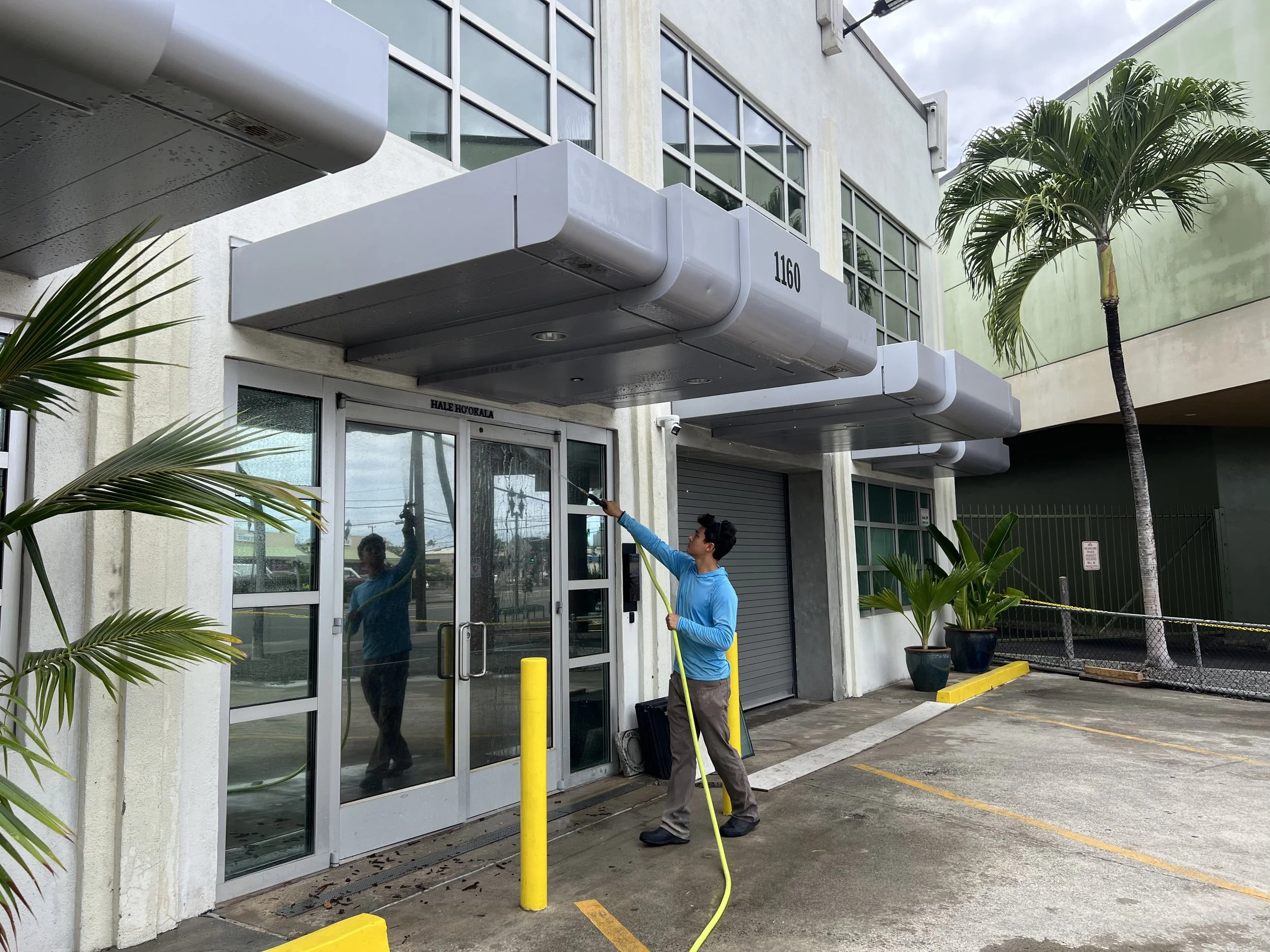 A man wearing a blue jacket and gray pants is cleaning the exterior glass door of a building using a spray and cloth. There are large potted plants and yellow parking barriers in the foreground. The building has large windows, white walls, and a gray awning with the number 1160. The sky is overcast.